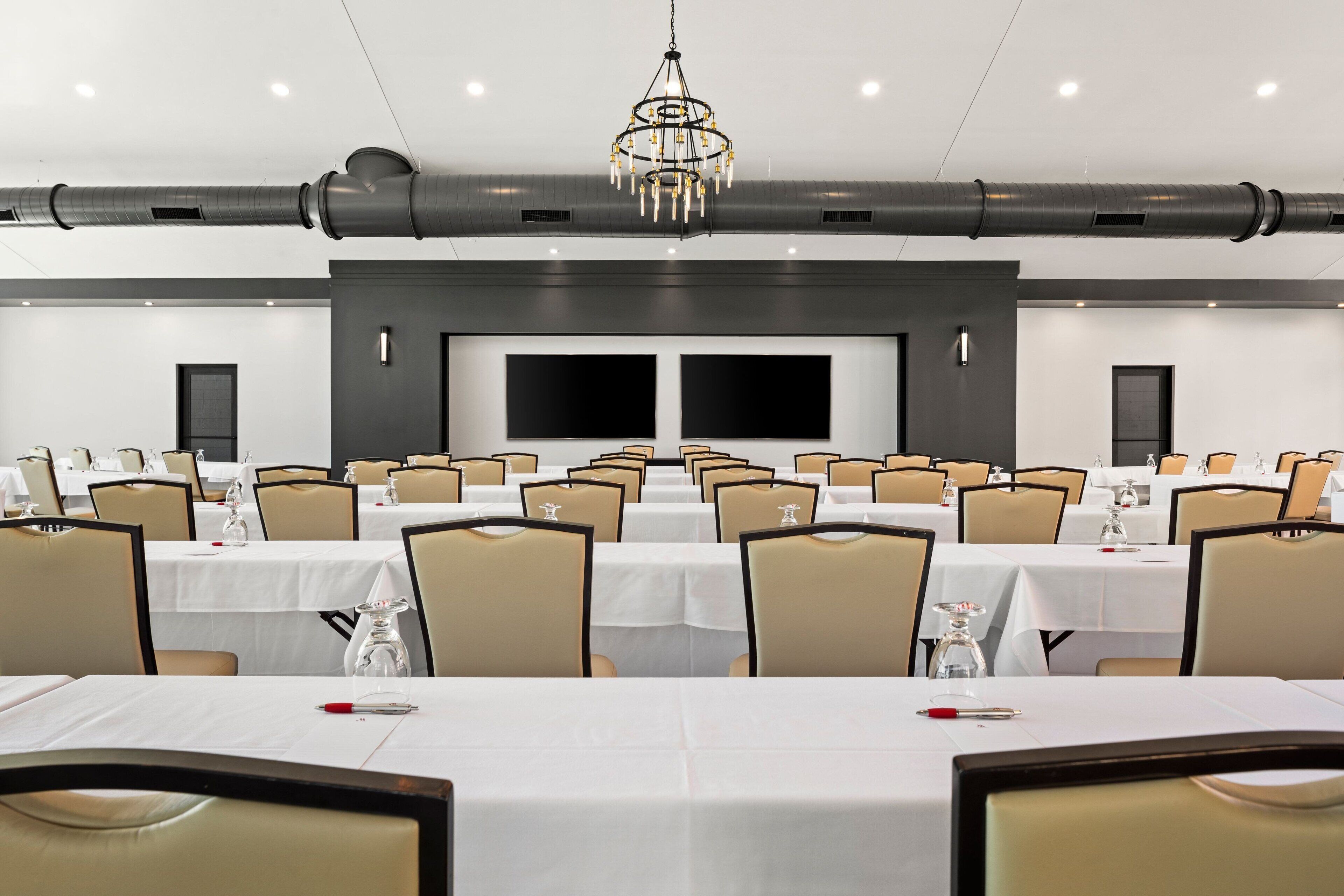 A conference room with multiple tables and chairs, large screens, and a chandelier.