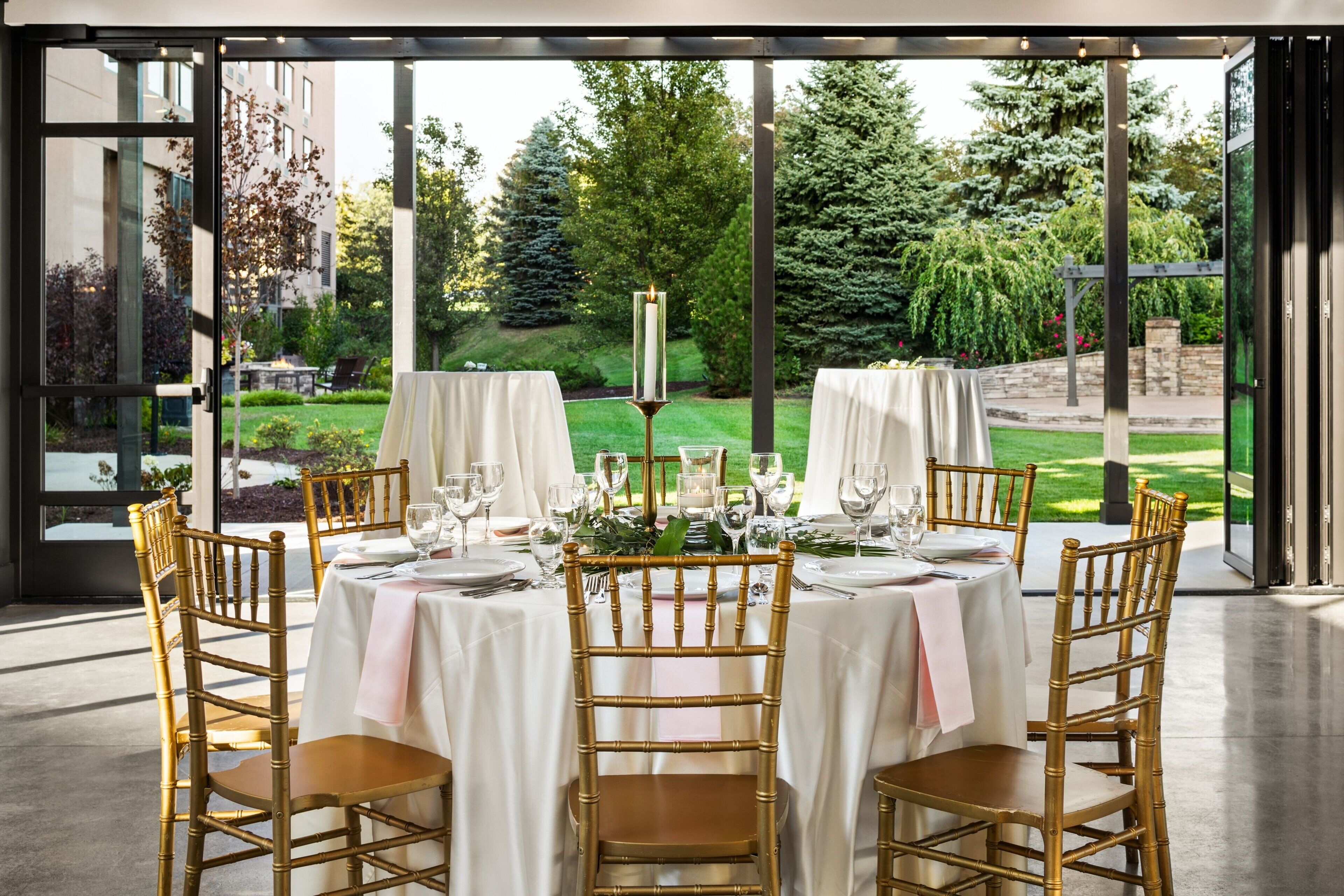 Comedor con mesas dispuestas para seis comensales, cada una con sillas doradas, manteles blancos y un centro de mesa con velas. La habitación tiene ventanas amplias que ofrecen vistas a un jardín cuidado.