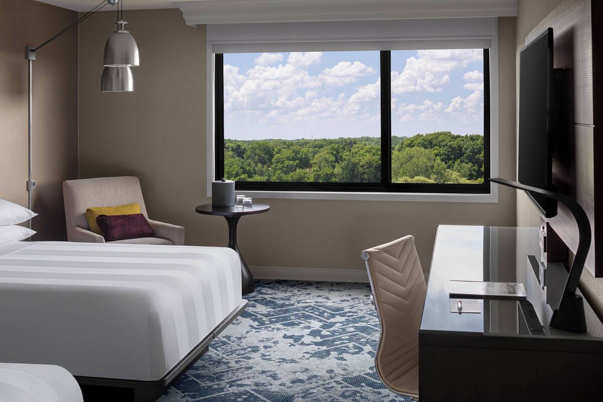 Une chambre d’hôtel équipée d’un lit, d’un bureau, d’une chaise, d’une télévision et d’une fenêtre donnant sur une forêt.