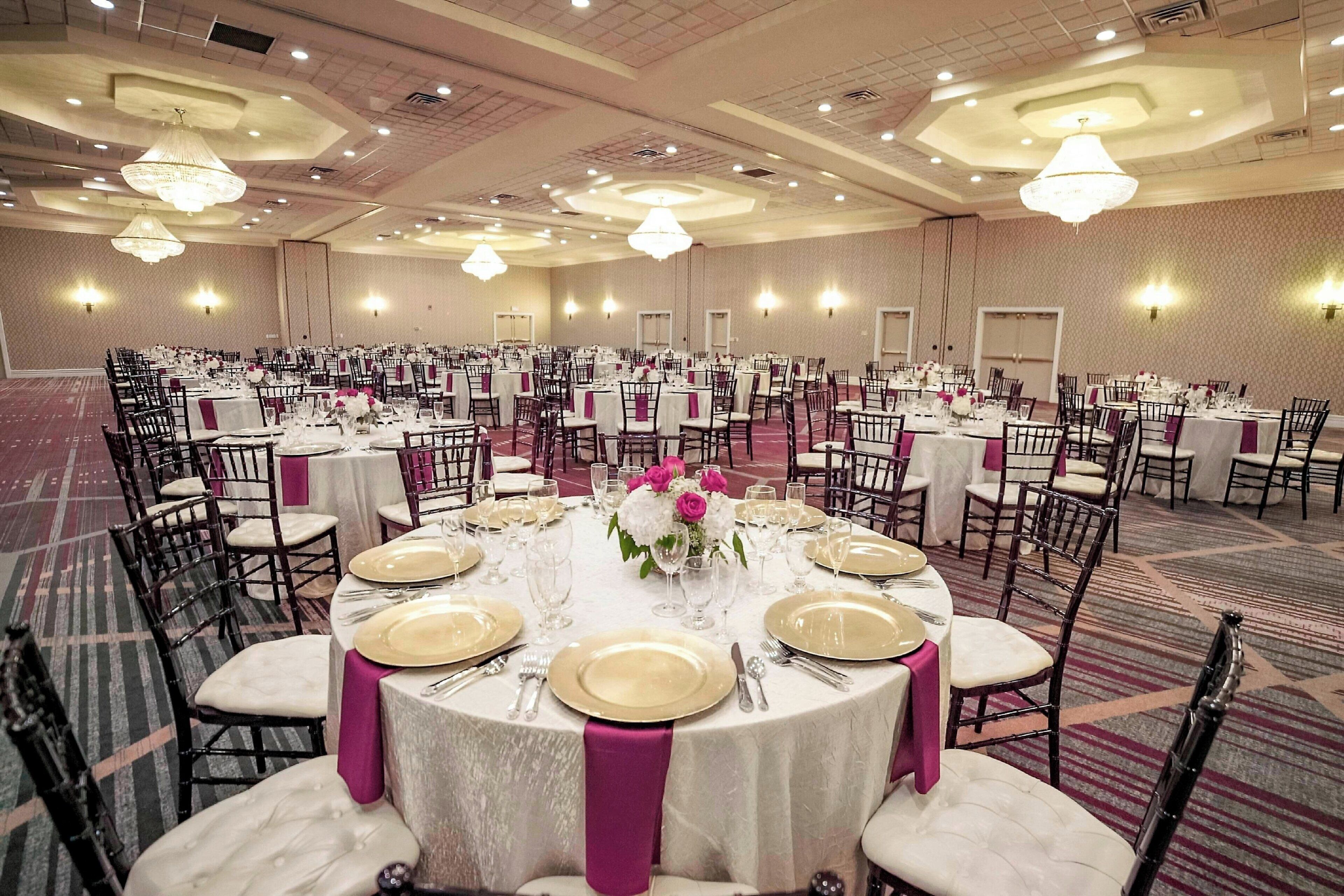 Une salle de banquet avec des tables rondes dressées pour un événement formel, agrémentée de nappes blanches, de assiettes décoratives dorées et de touches de rose.