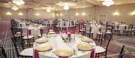 Une salle de banquet avec des tables rondes dressées pour un événement formel, agrémentée de nappes blanches, de assiettes décoratives dorées et de touches de rose.