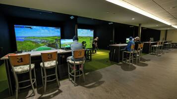 A room with multiple screens displaying a golf game, chairs with numbered backrests, and a green carpeted floor.