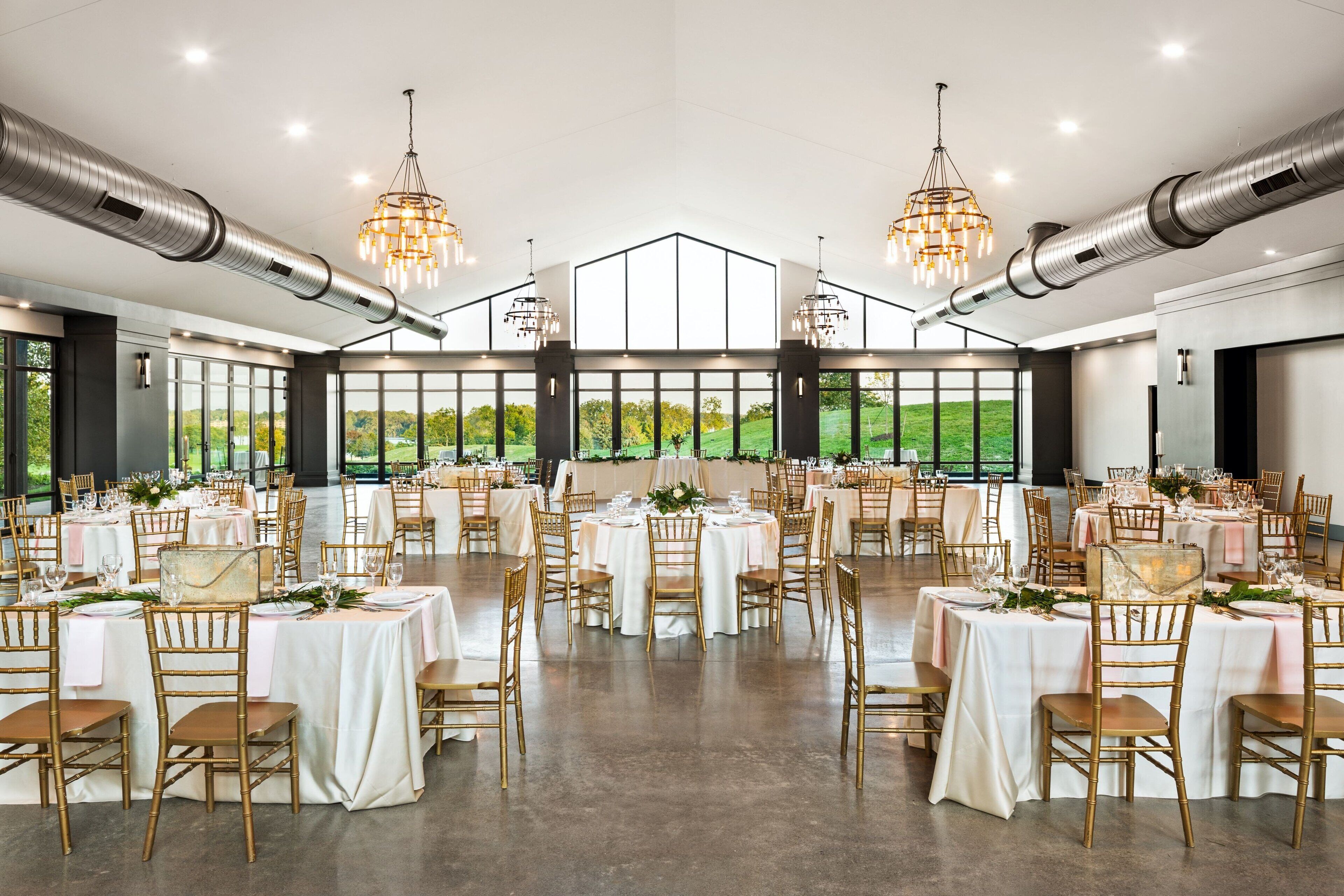 A spacious banquet hall with multiple round tables, gold chairs, and large windows offering a view of the outdoors.