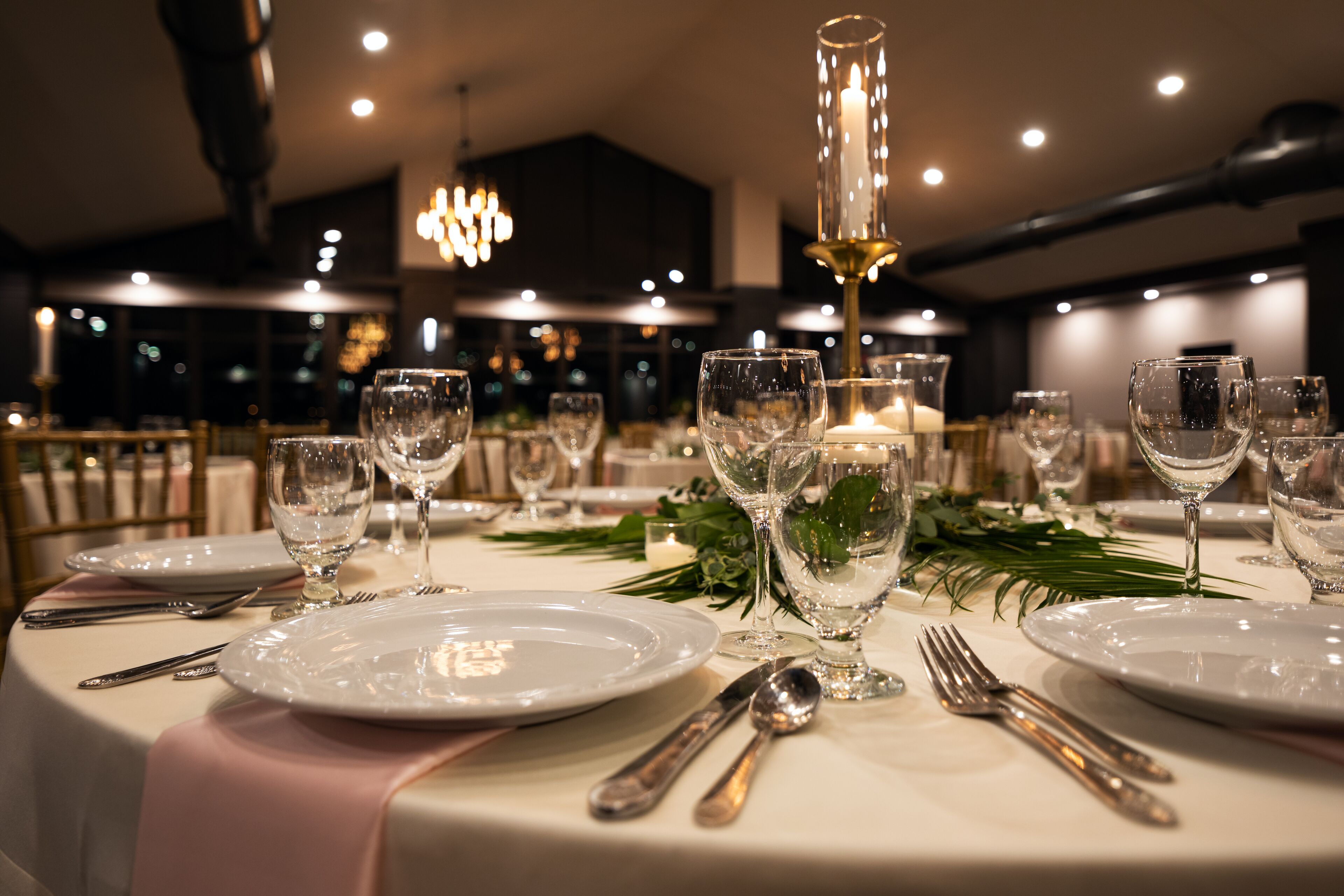 Una mesa de comedor elegantemente dispuesta con platos, cubiertos y copas de vino, decorada con follaje y un centro de mesa con velas.