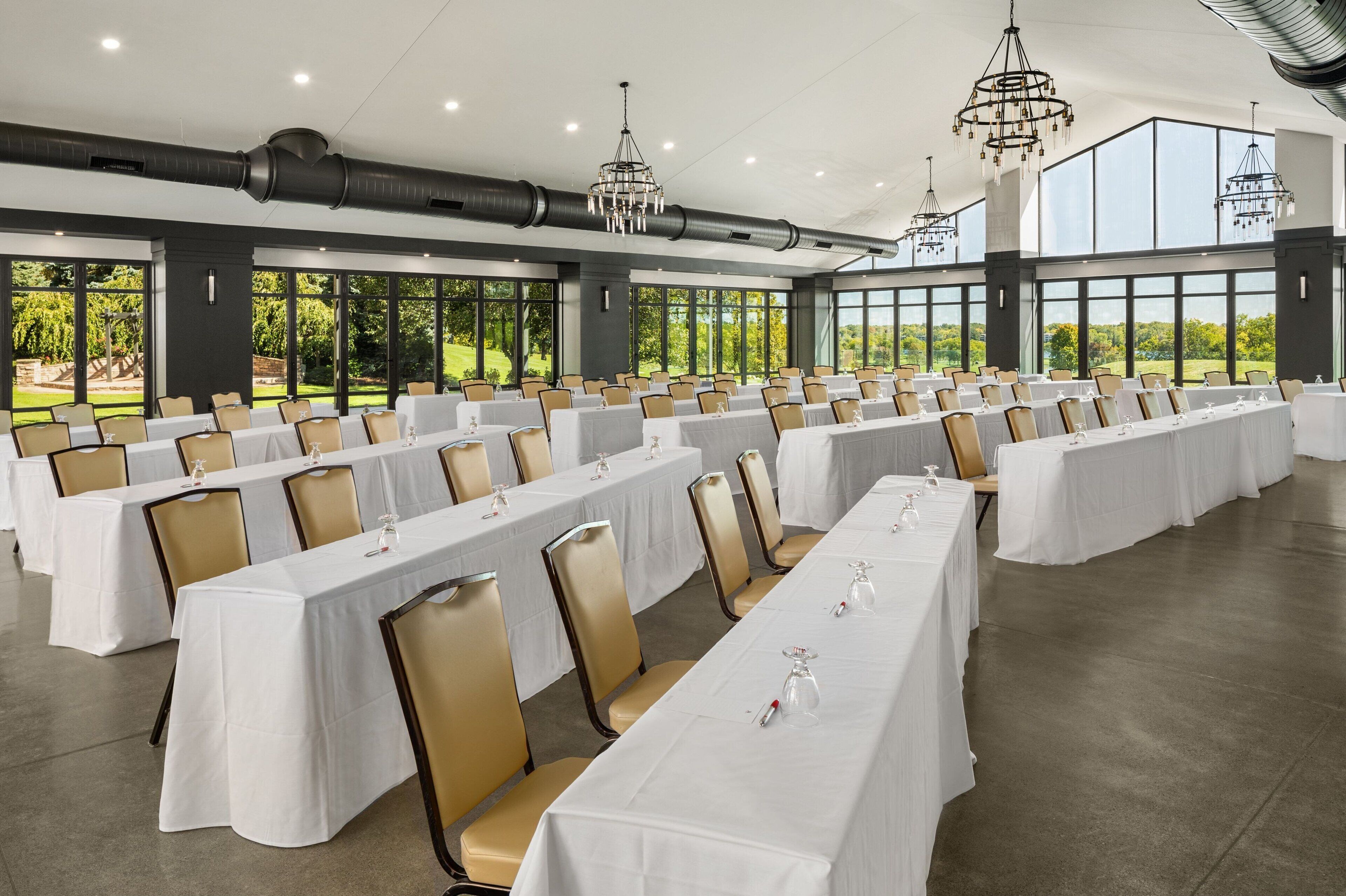 A large conference room with rows of tables and chairs, large windows, and modern chandeliers.
