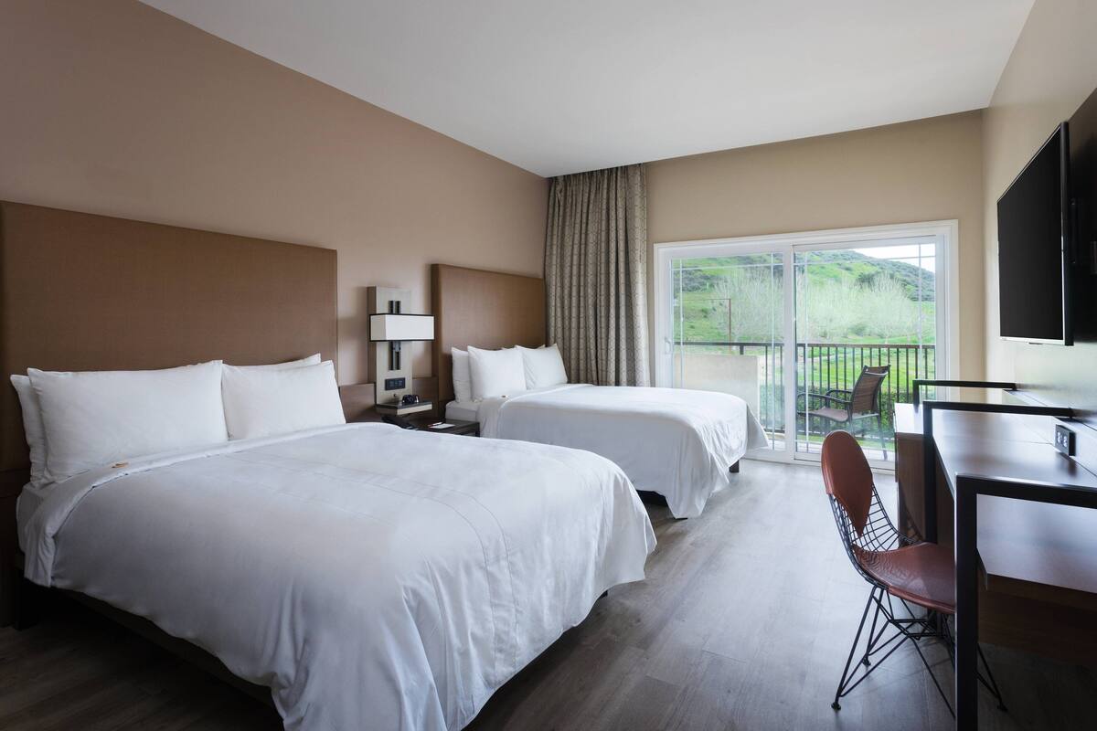 A hotel room with two beds, a desk, a chair, and a balcony with a view of greenery.