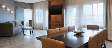 A modern living room with a dining area, a flat-screen TV mounted on a wooden panel, and a large window with curtains.