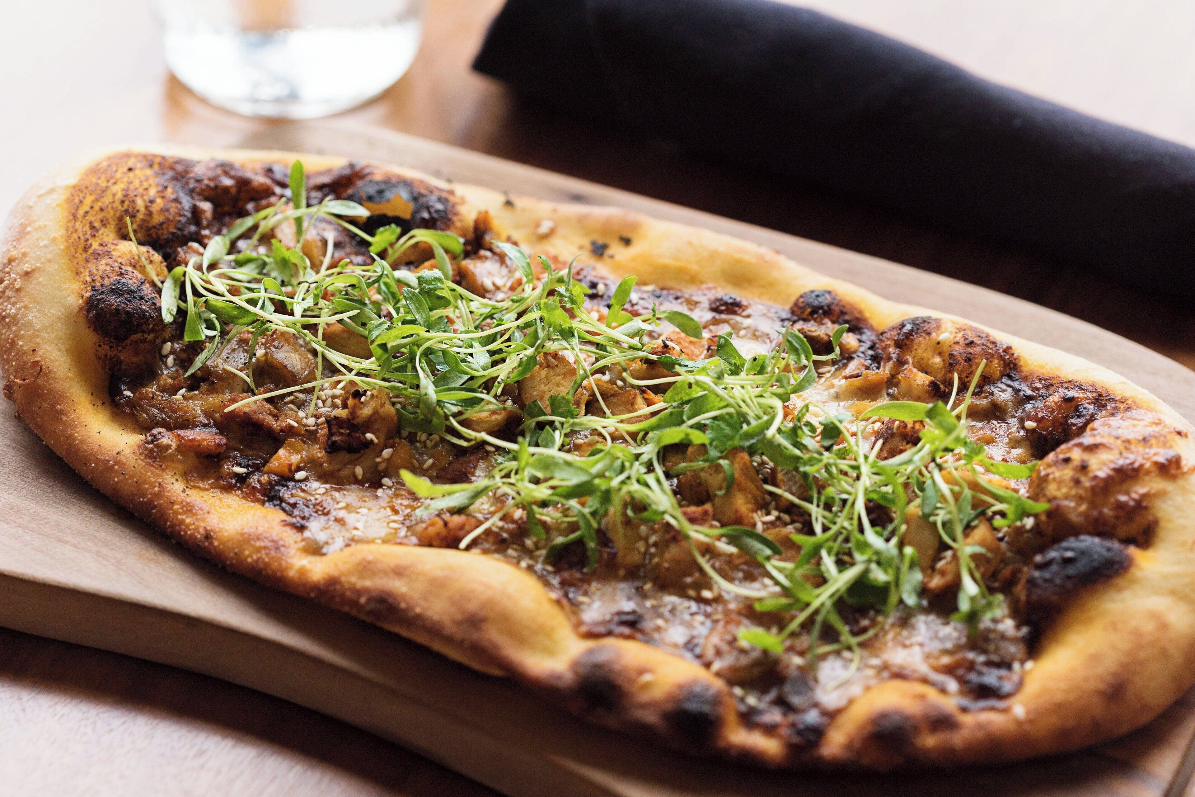 Pizza sobre tabla de madera con bordes quemados, adornada con vegetales verdes, sobre una mesa con servilleta negra y un vaso de agua al fondo.