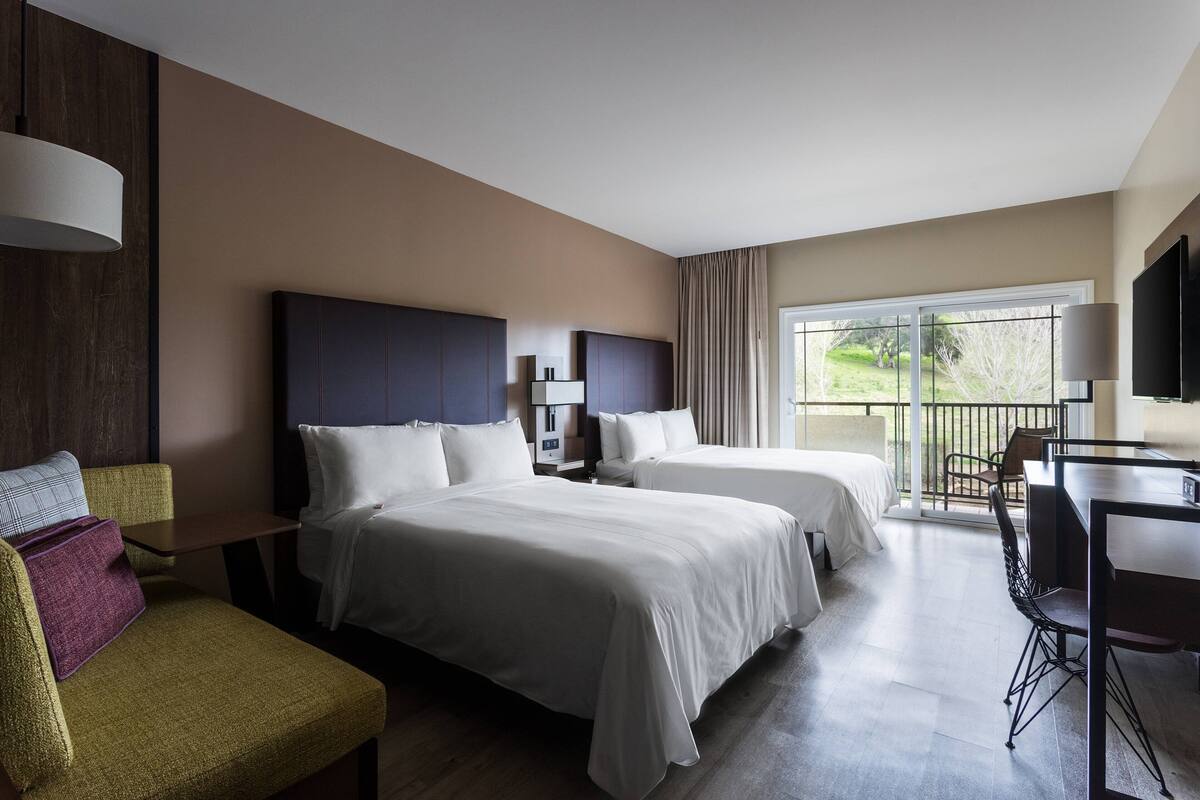 A hotel room with two beds, a desk, a chair, and a balcony with a view.