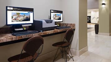 Un comptoir d’accueil avec deux écrans d’ordinateur affichant le site web de l’hôtel, une imprimante, un clavier et deux chaises au design minimaliste.