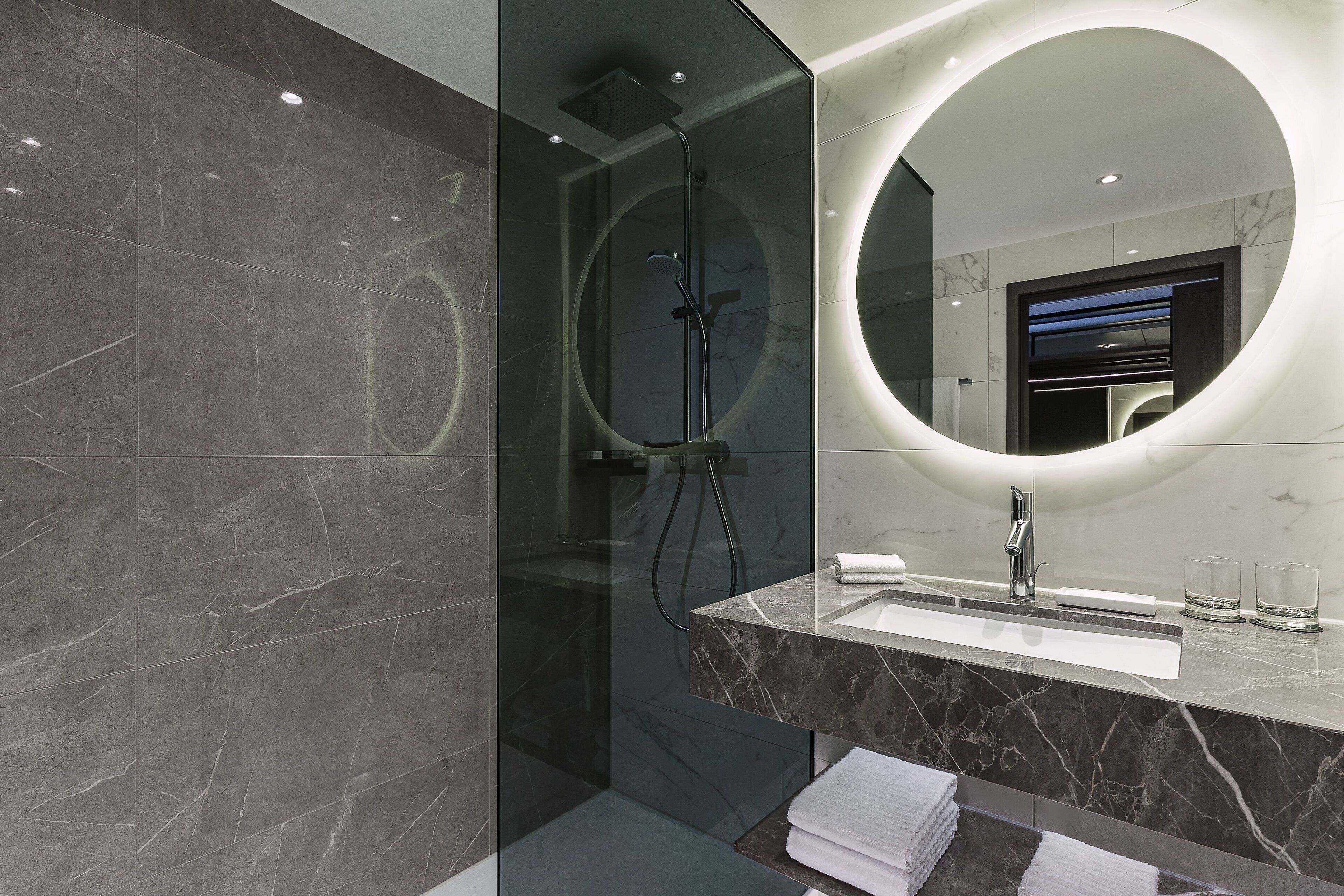 A modern bathroom with a glass-enclosed shower, a marble vanity, and a large mirror with a circular light.