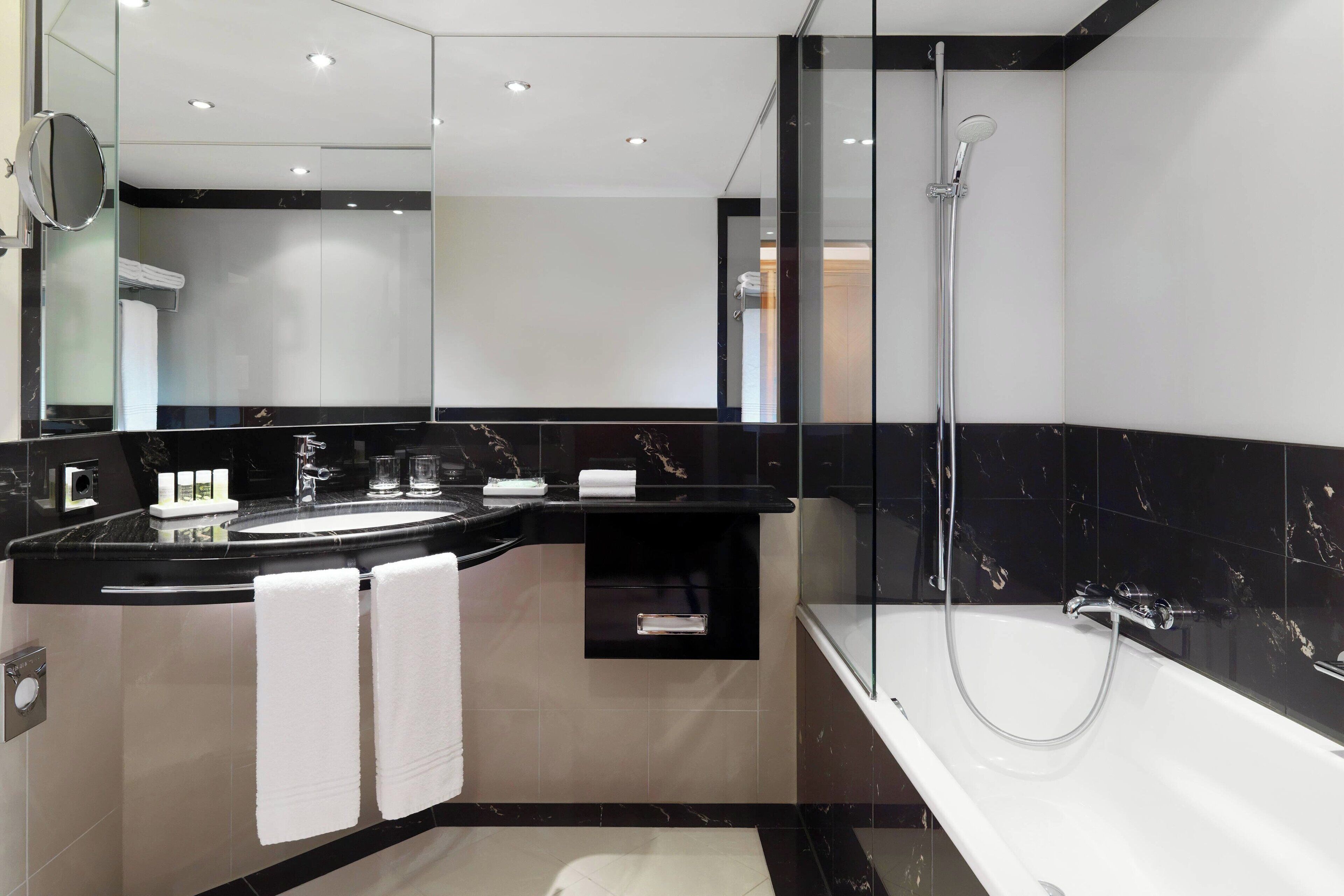 Un baño moderno con diseño de mármol blanco y negro, cabina de ducha con mampara de vidrio y tocador con lavamanos y toallero.