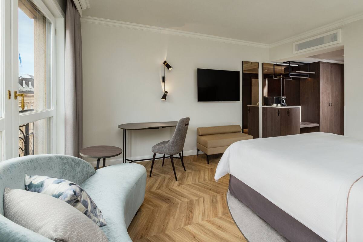 A hotel room with a bed, a sofa, a desk, a chair, a TV, and a wardrobe.