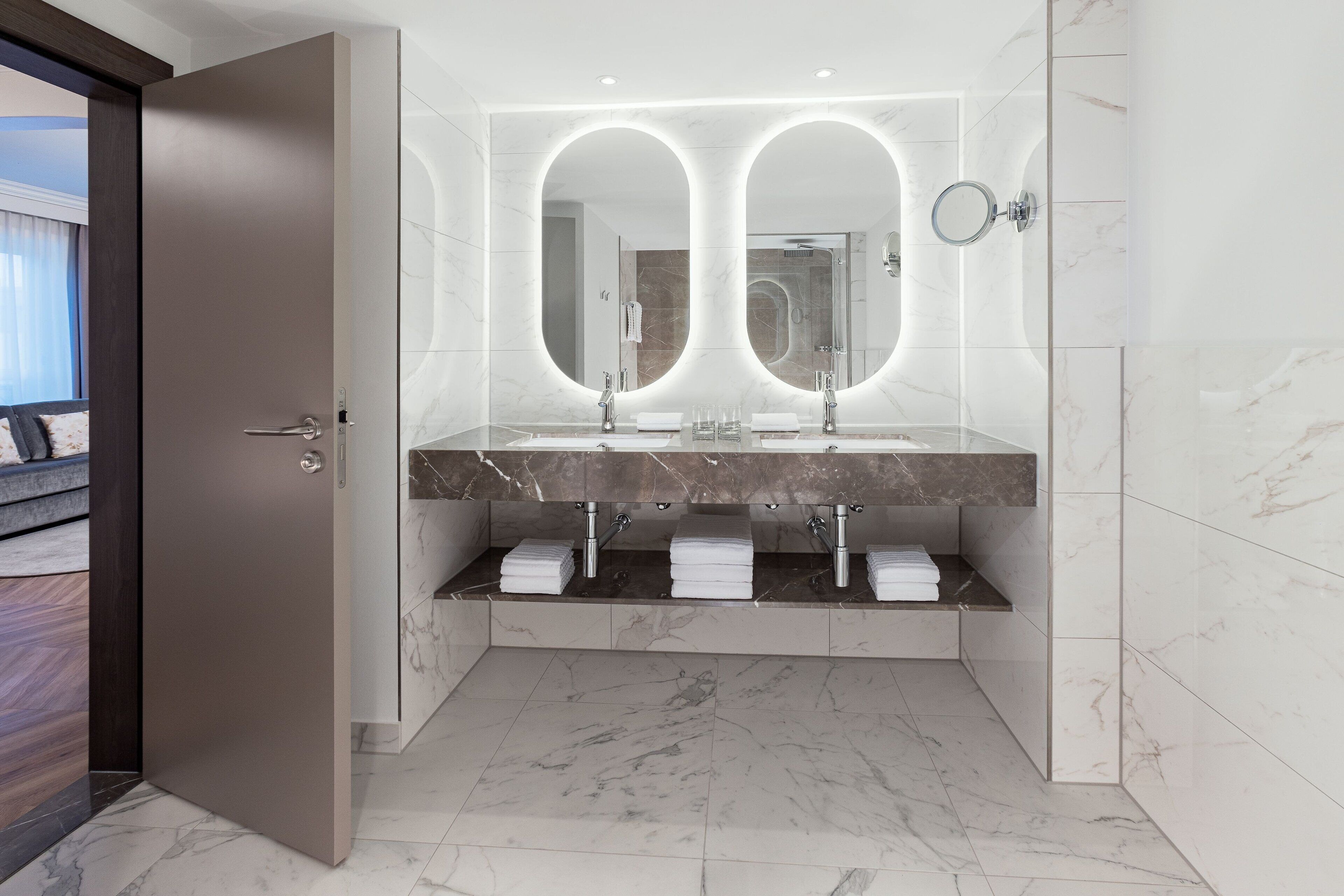 Ein modernes Badezimmer mit Doppelwaschbecken, Marmorboden und einem großen Spiegel mit integrierter Beleuchtung.