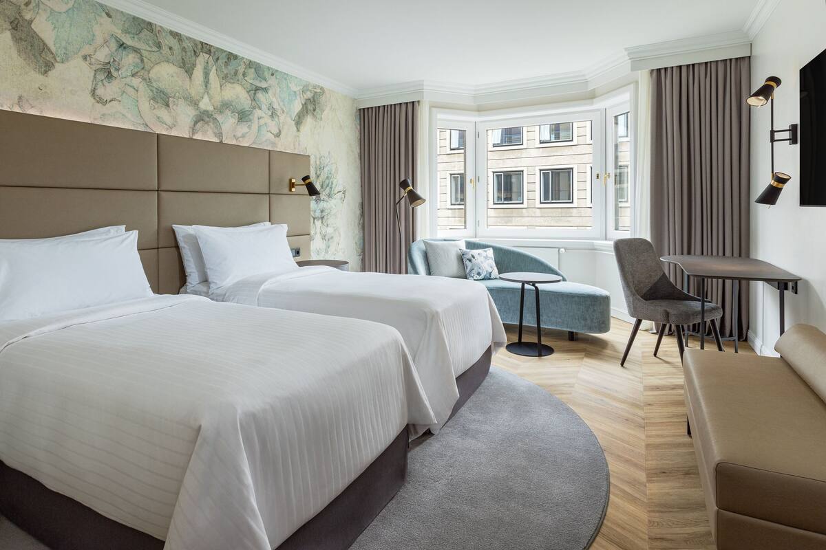 A hotel room with two beds, a sofa, a chair, a small table, and a view of buildings through a window.