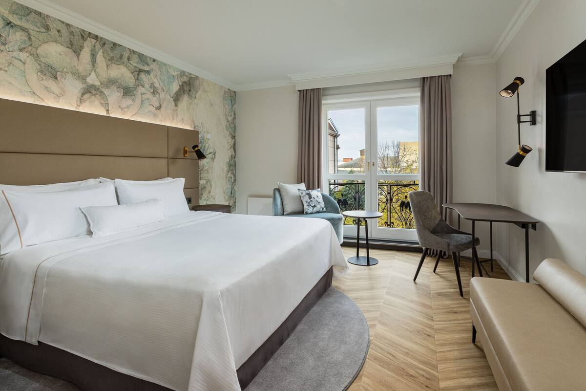 A hotel room with a large bed, a seating area with a table and chairs, a balcony with a view, and a mounted TV.