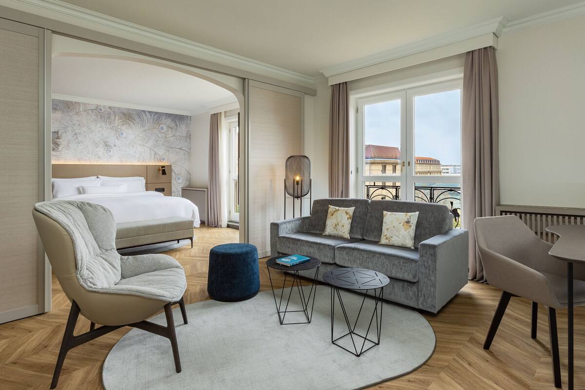 A hotel room with a bed, a sofa, a chair, a small table, and a large window with a view of buildings.