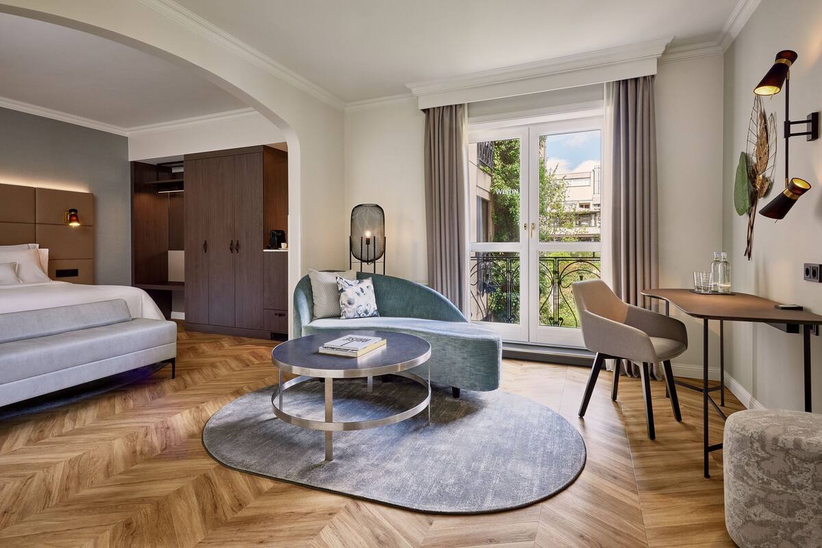 A modern hotel room with a bed, sofa, round coffee table, and a balcony with curtains.