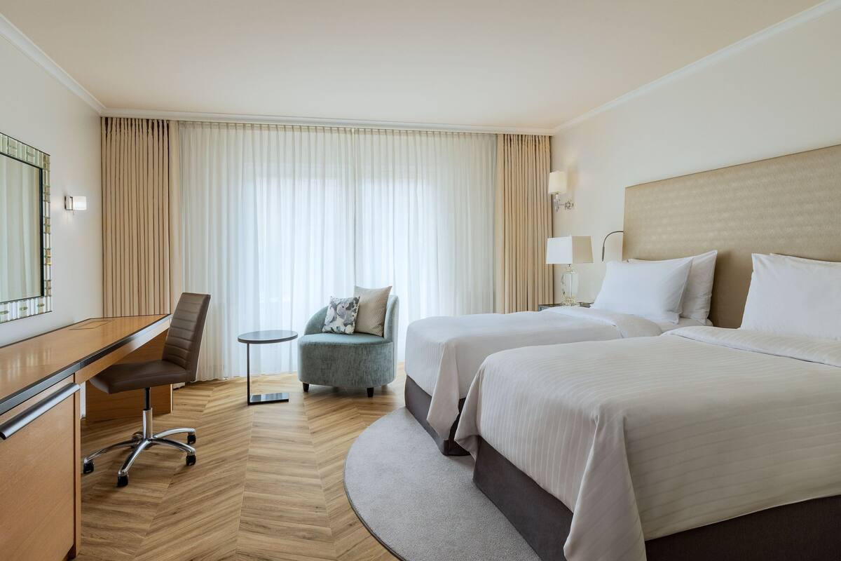 A hotel room with a desk, two beds, a chair, and a large window with curtains.