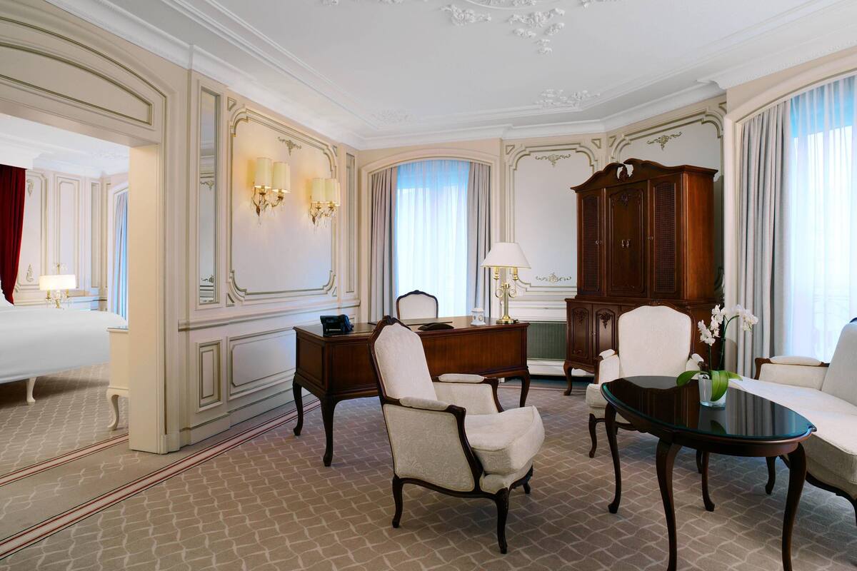 A hotel room with a bed, two armchairs, a desk, a large wooden cabinet, and a glass coffee table.