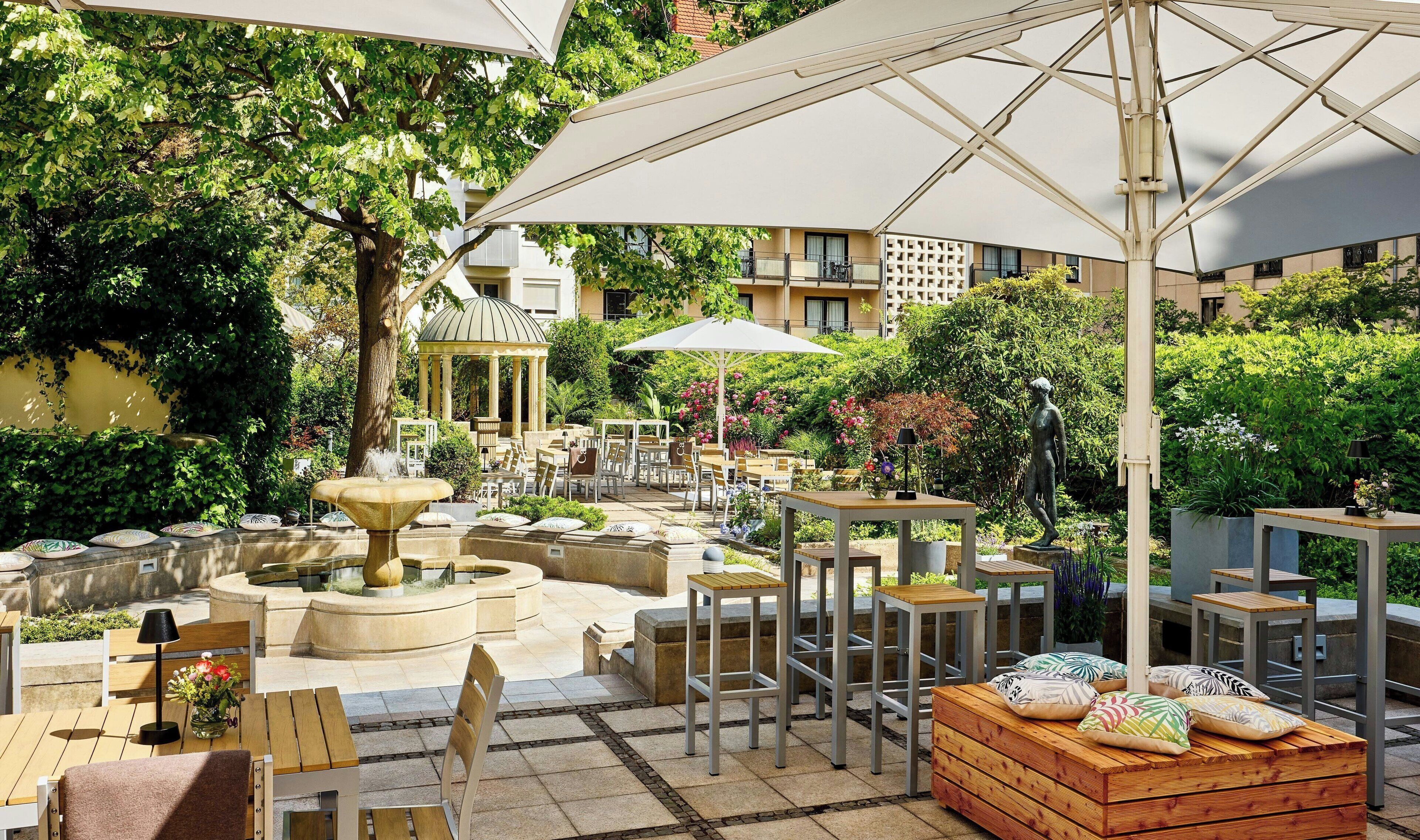 En udendørs terrasse med borde, stole, parasoller og en springvand.