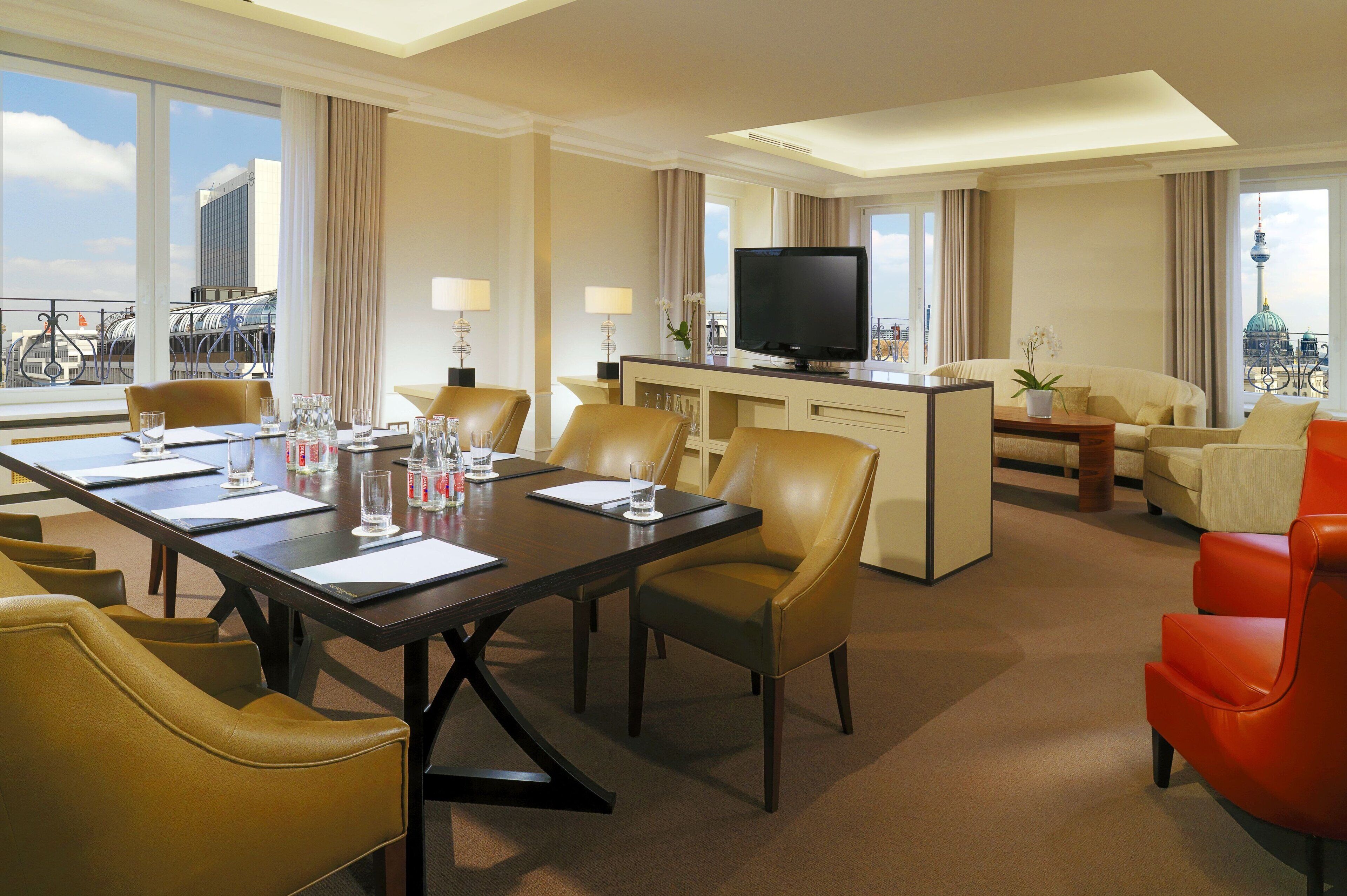 A hotel room with a dining area, a living area with a sofa and armchairs, a television, and a view of a cityscape.