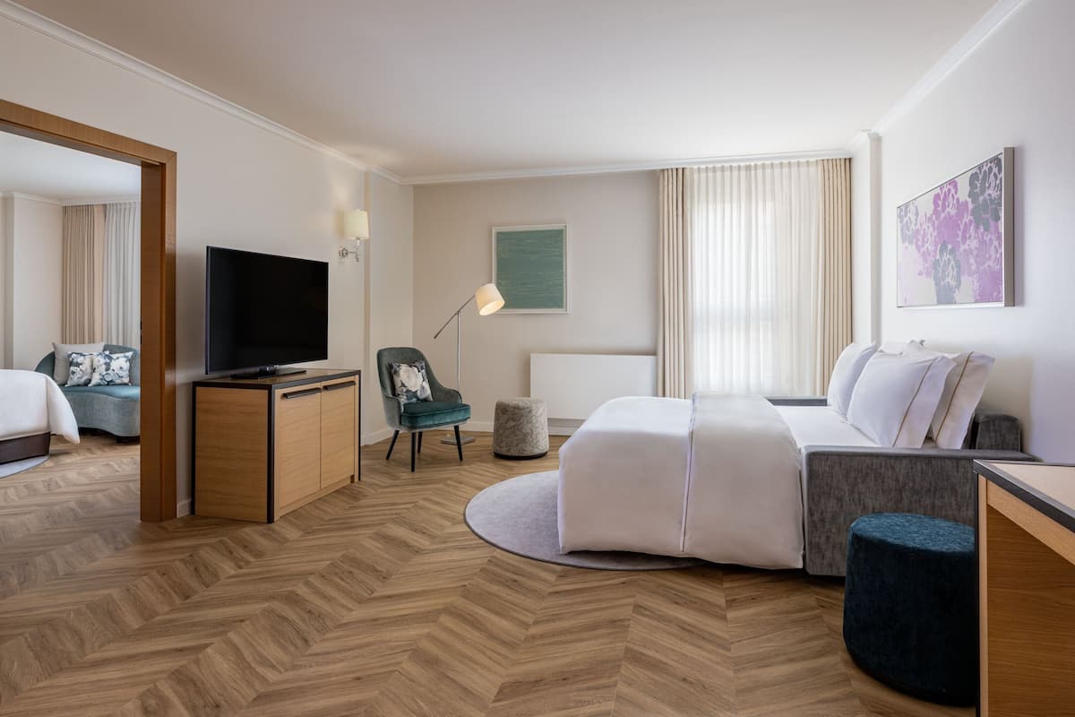 A hotel room with a television, sofa, ottoman, and a desk with a lamp.