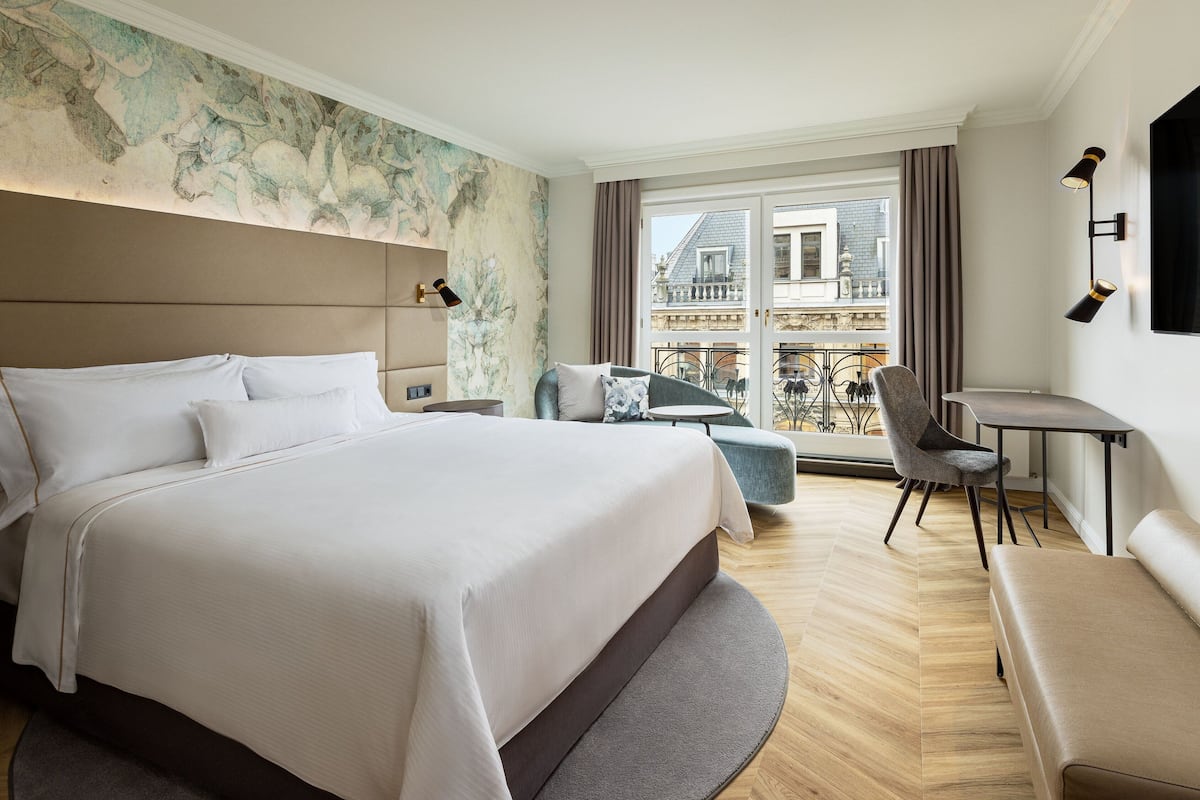 A modern hotel room with a large bed, a small round table, a chair, and a balcony with a view of the city.