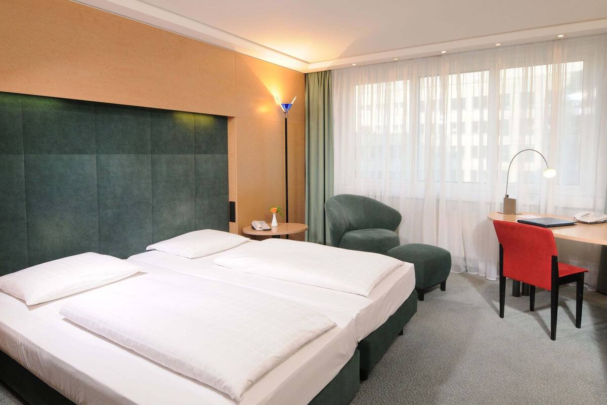 Ein Hotelzimmer mit einem großen Bett, einem Schreibtisch mit Stuhl, einer Lampe und einem Fenster mit Vorhängen.