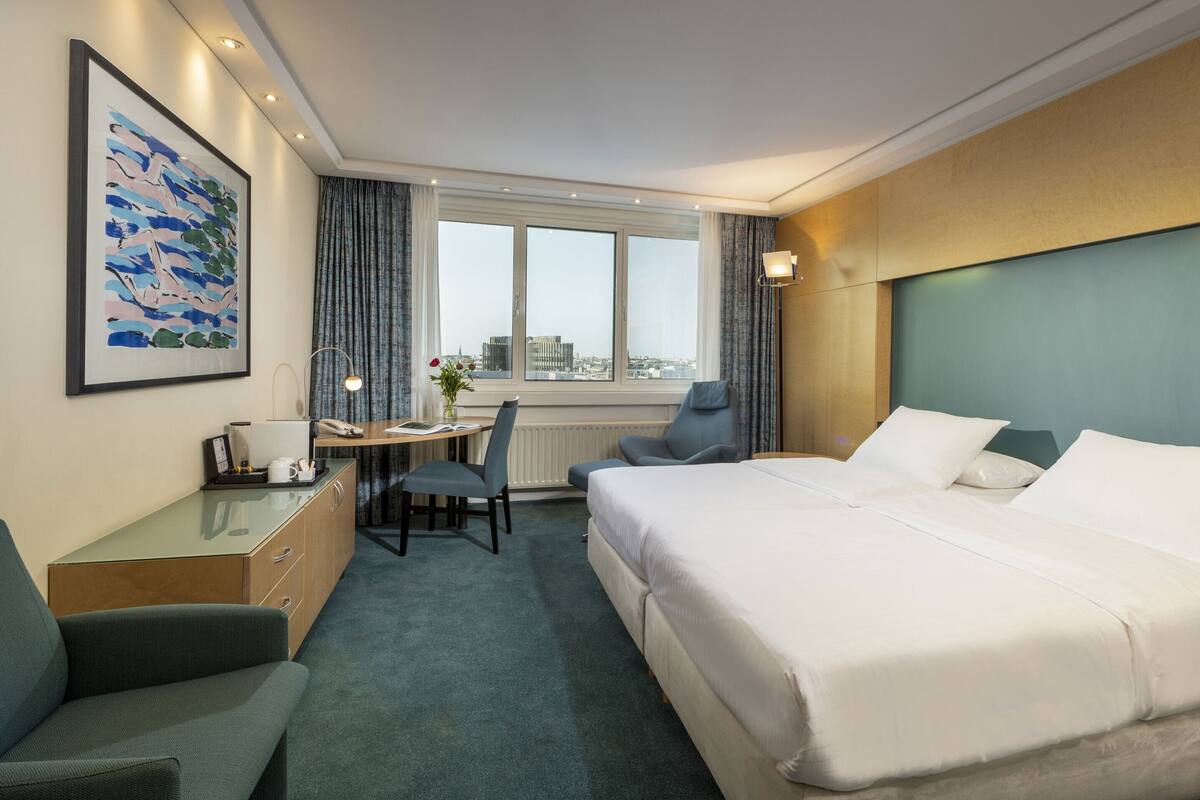 Ein Hotelzimmer mit einem großen Bett, einem Schreibtisch mit Stuhl, einer kleinen Couch, einem Fenster mit Vorhängen und einem gerahmten Bild an der Wand.