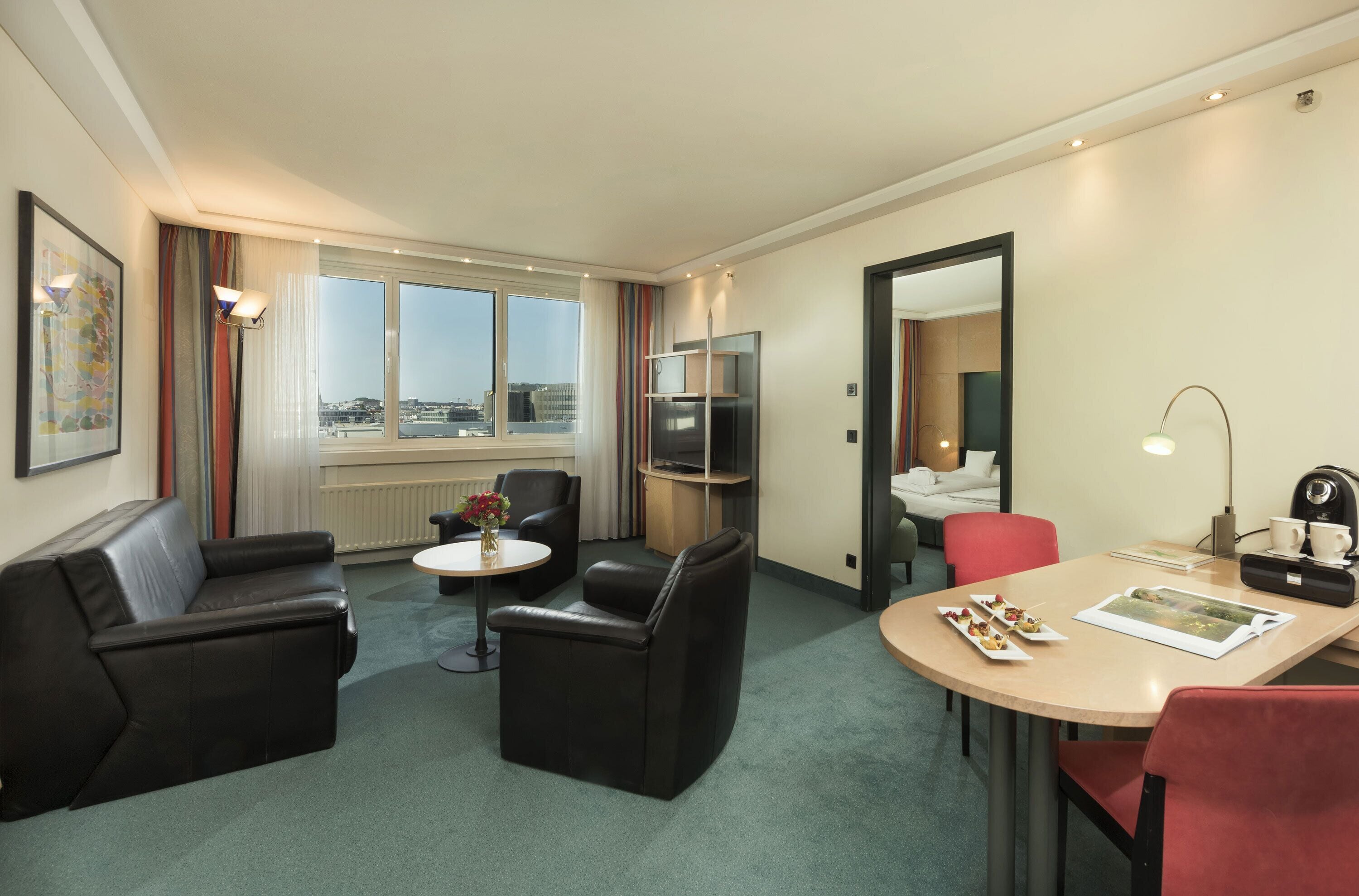 Habitación de hotel moderna con sofá, sillas, una mesa de comedor, televisor y un amplio ventanal con vistas a la ciudad.