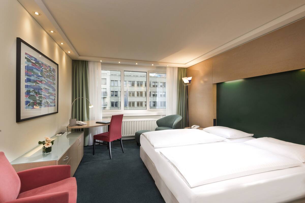 Ein Hotelzimmer mit Bett, Schreibtisch, Stuhl und einem Fenster mit Blick auf Gebäude.