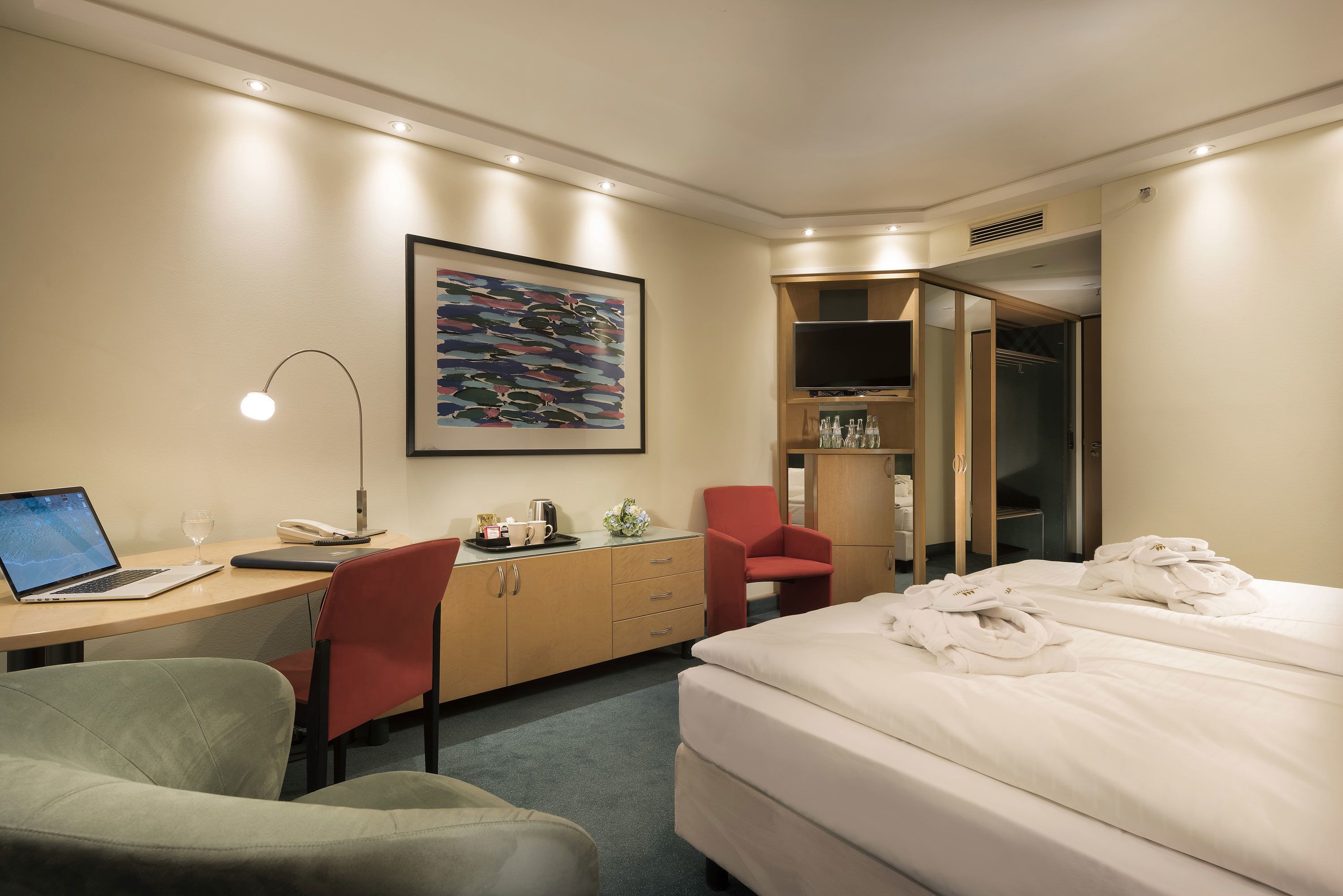 A hotel room with a bed, a desk with a laptop, a chair, a red chair, a TV, and a painting on the wall.