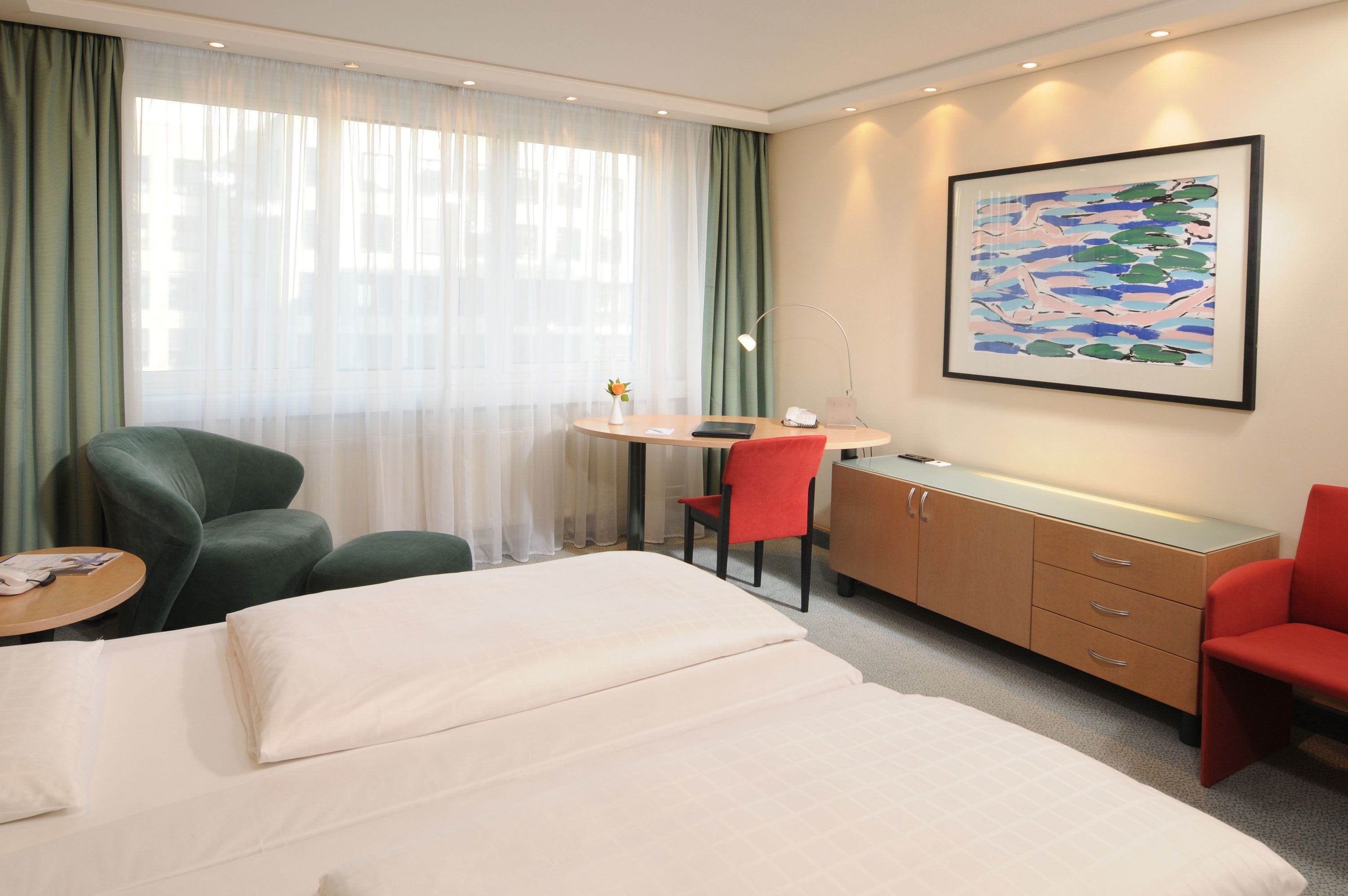 A hotel room with a bed, two chairs, a desk, a lamp, and a painting on the wall.