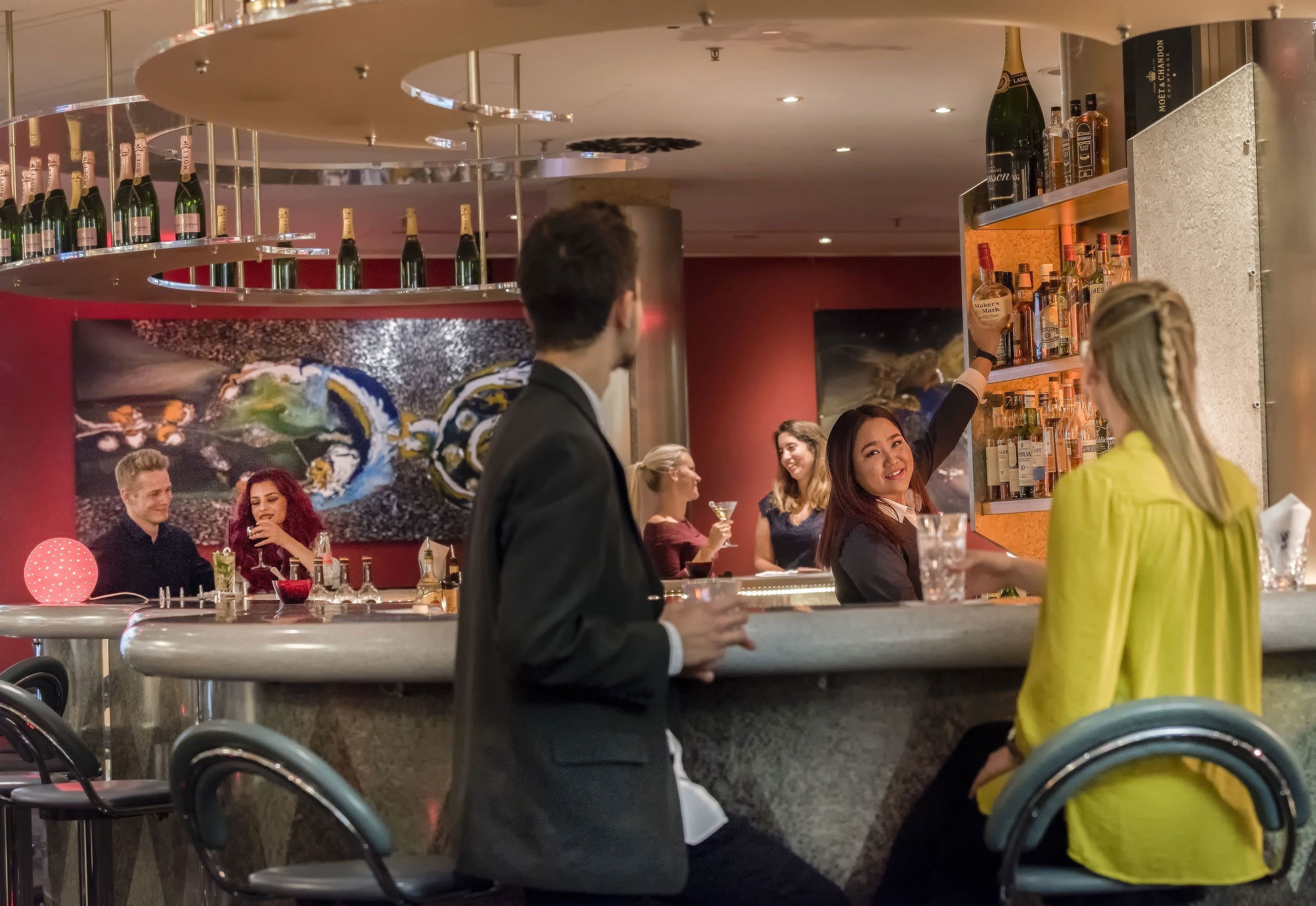 A bar area with a circular chandelier, a large decorative painting, and a variety of bottles on shelves.