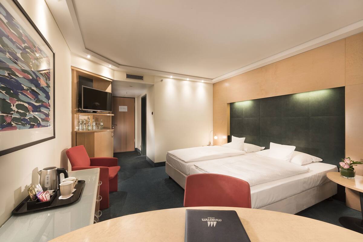 Ein Hotelzimmer mit einem Bett, einem Schreibtisch mit Frühstücksgegenständen und einem roten Sessel sowie einem Fernseher.