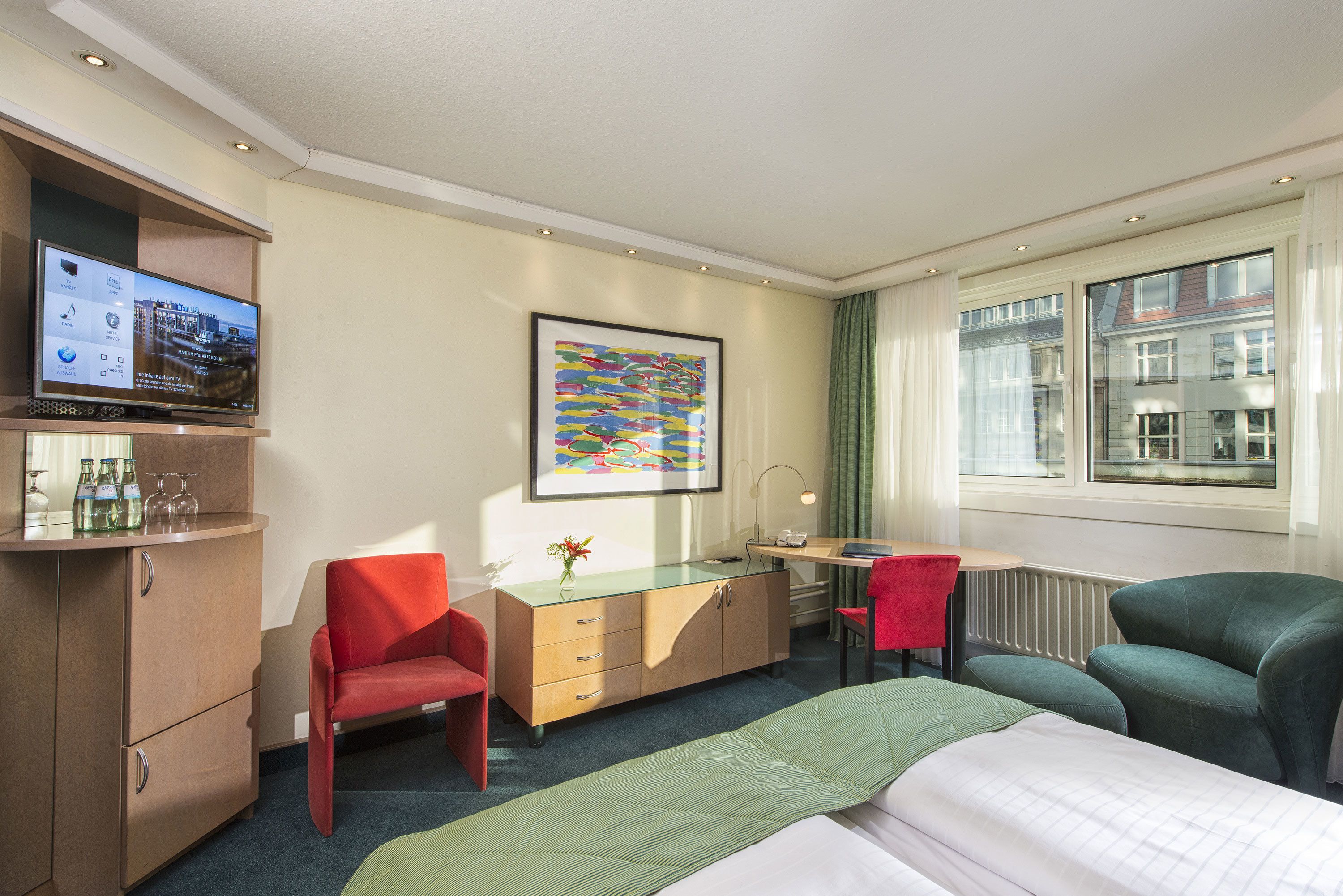 A hotel room with a bed, a desk, a TV, a red chair, a green chair, and a framed picture on the wall.
