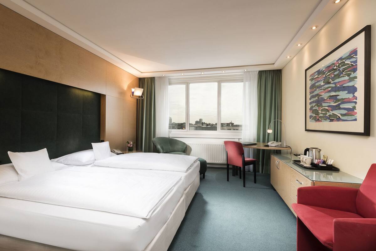 Ein Hotelzimmer mit einem großen Bett, einem Schreibtisch, einem Sessel und Blick auf die Stadt durch das Fenster.