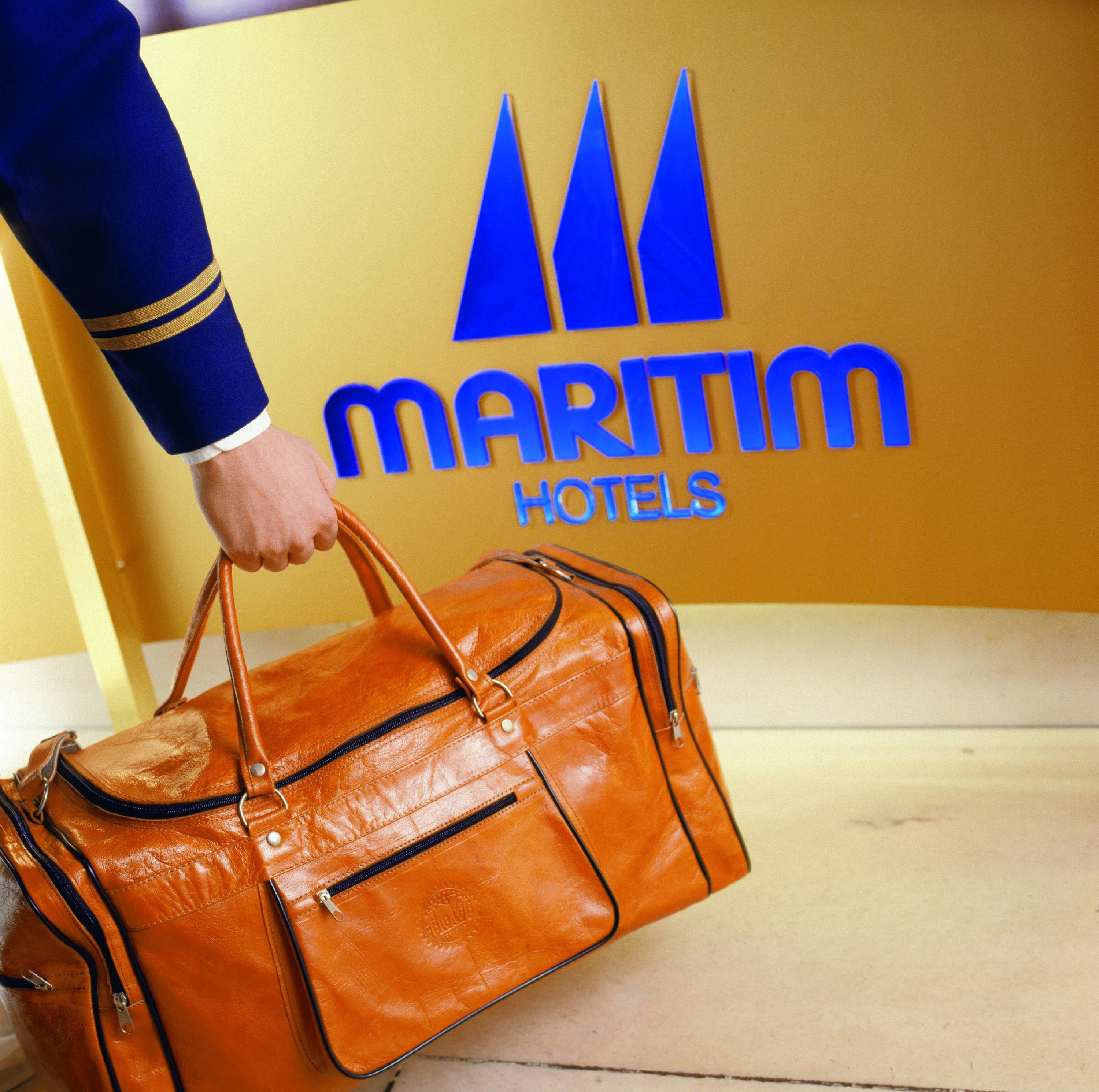 Una mano sostiene un bolso de cuero marrón frente a una pared amarilla con el texto "MARITIM HOTELS".