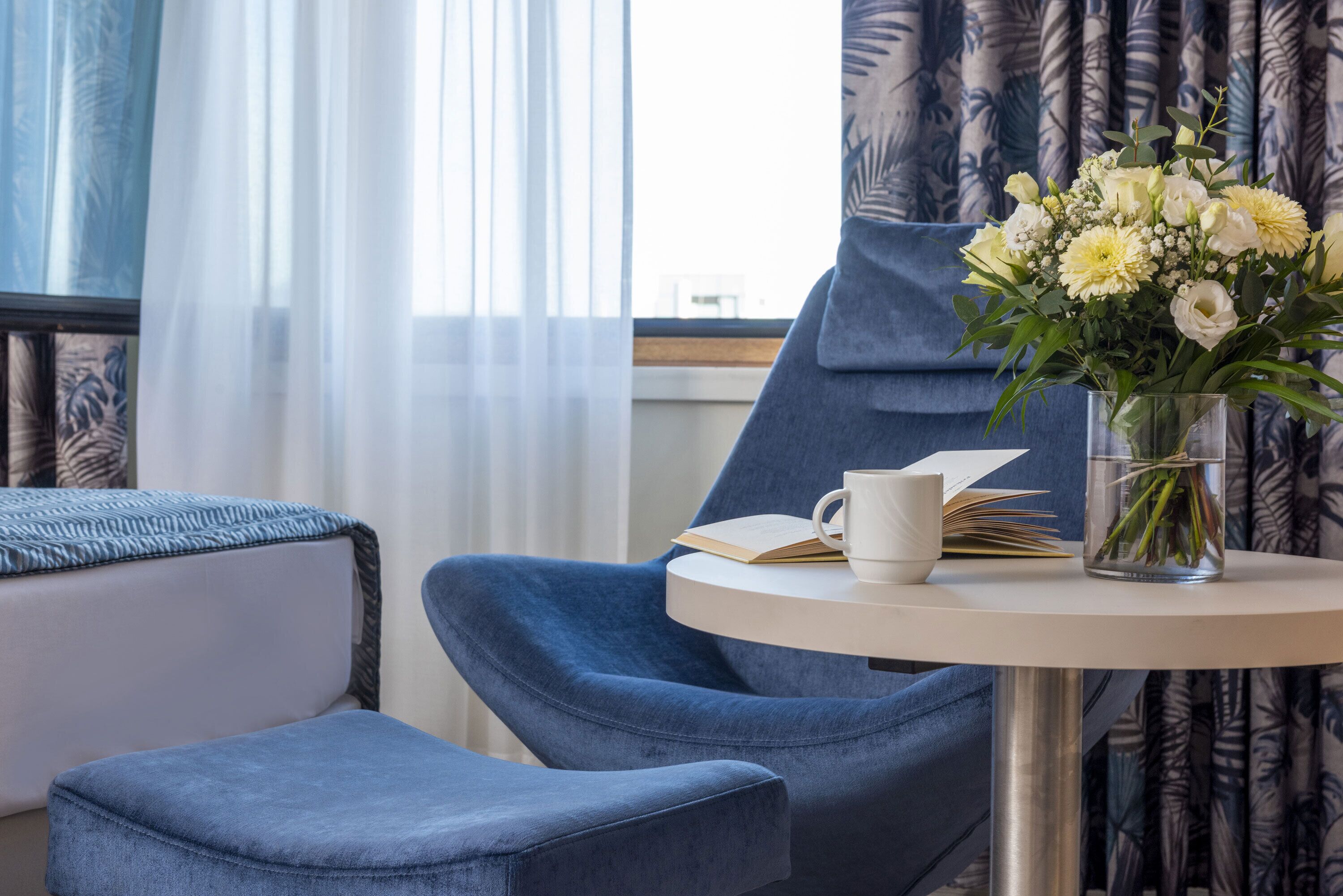 A room with a blue armchair, a small round table with a book, a white mug, and a vase with flowers.