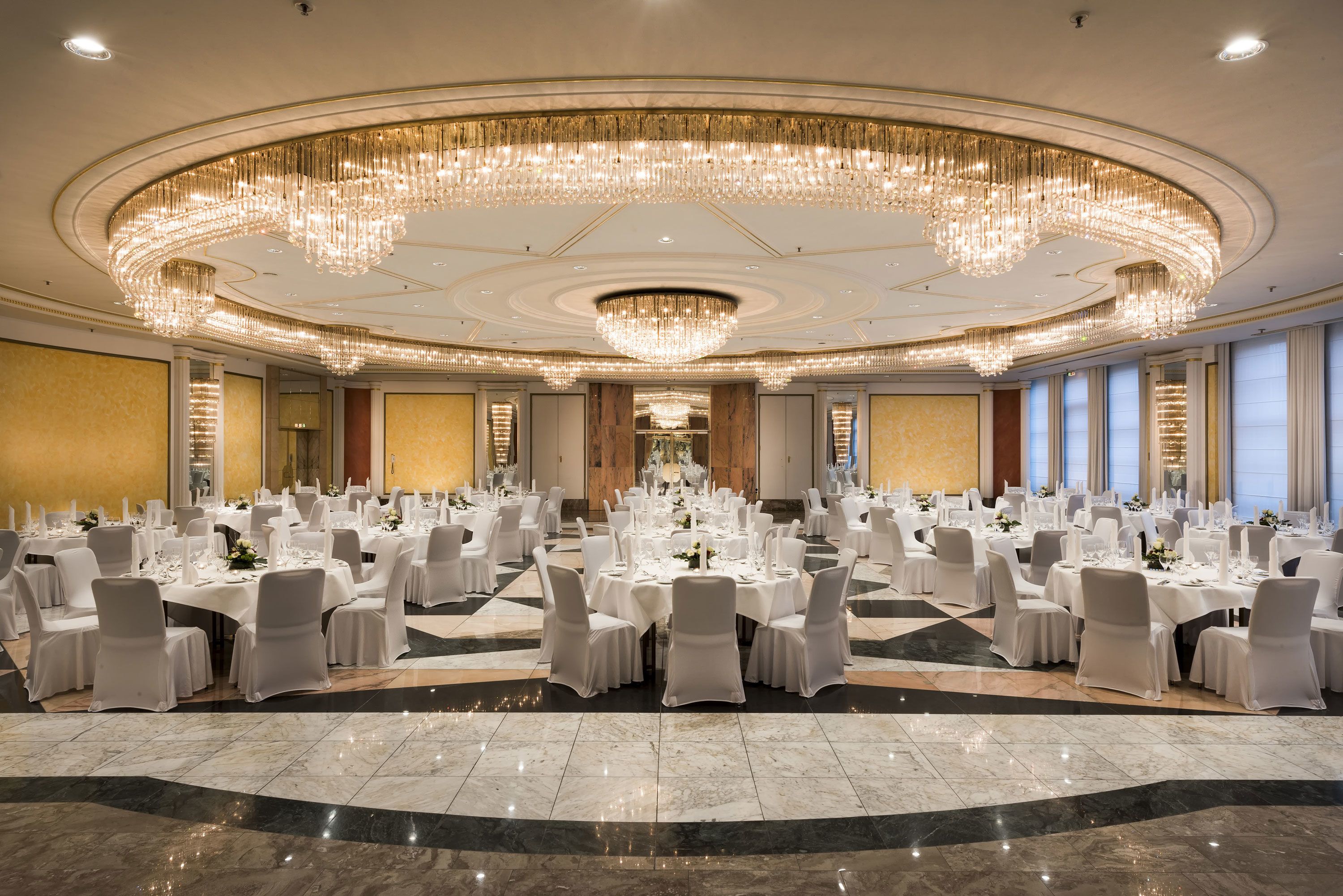 A large banquet hall with round tables, white chairs, and a grand chandelier.