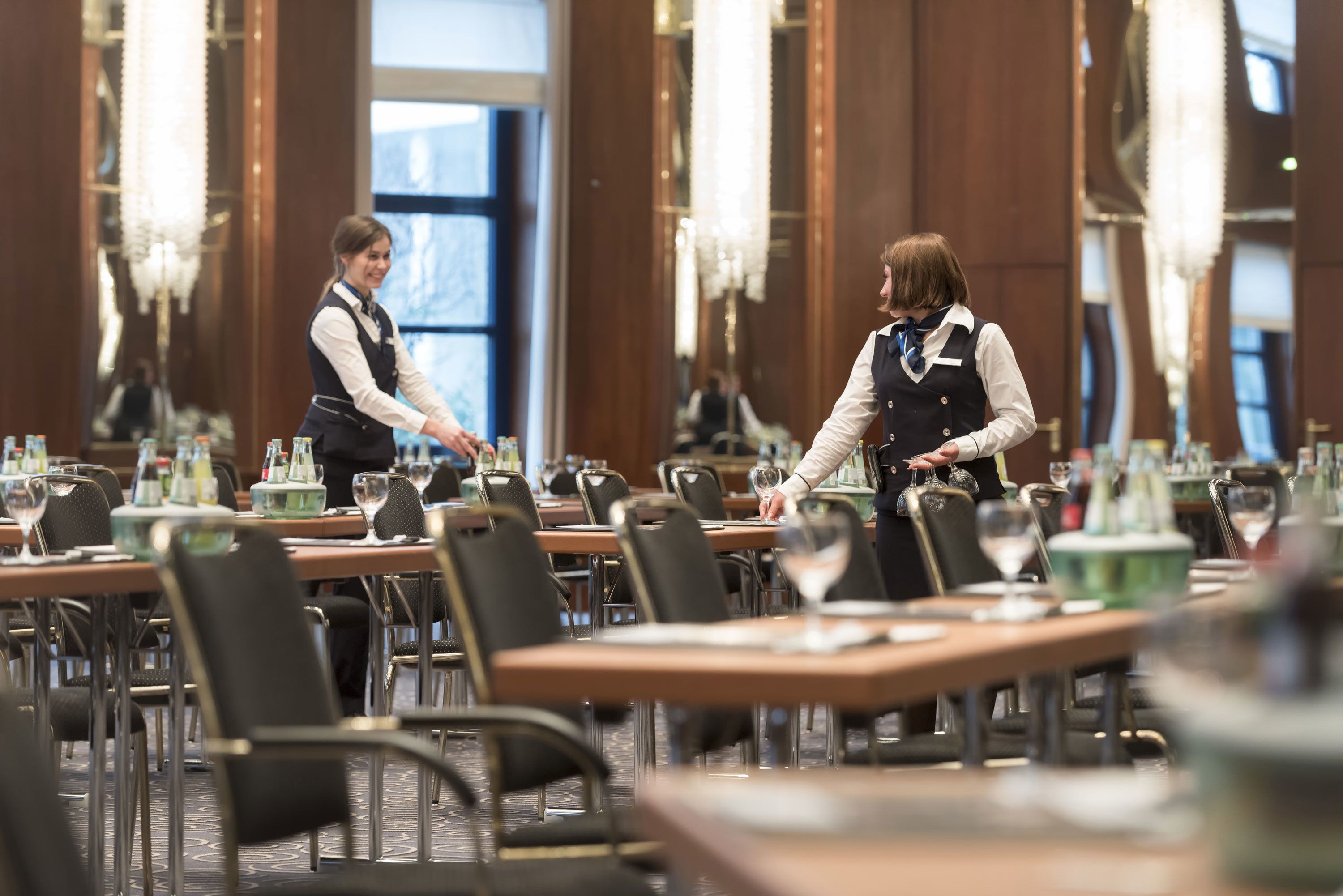 A formal dining area with tables set for service, waitstaff in uniform, and chandeliers.