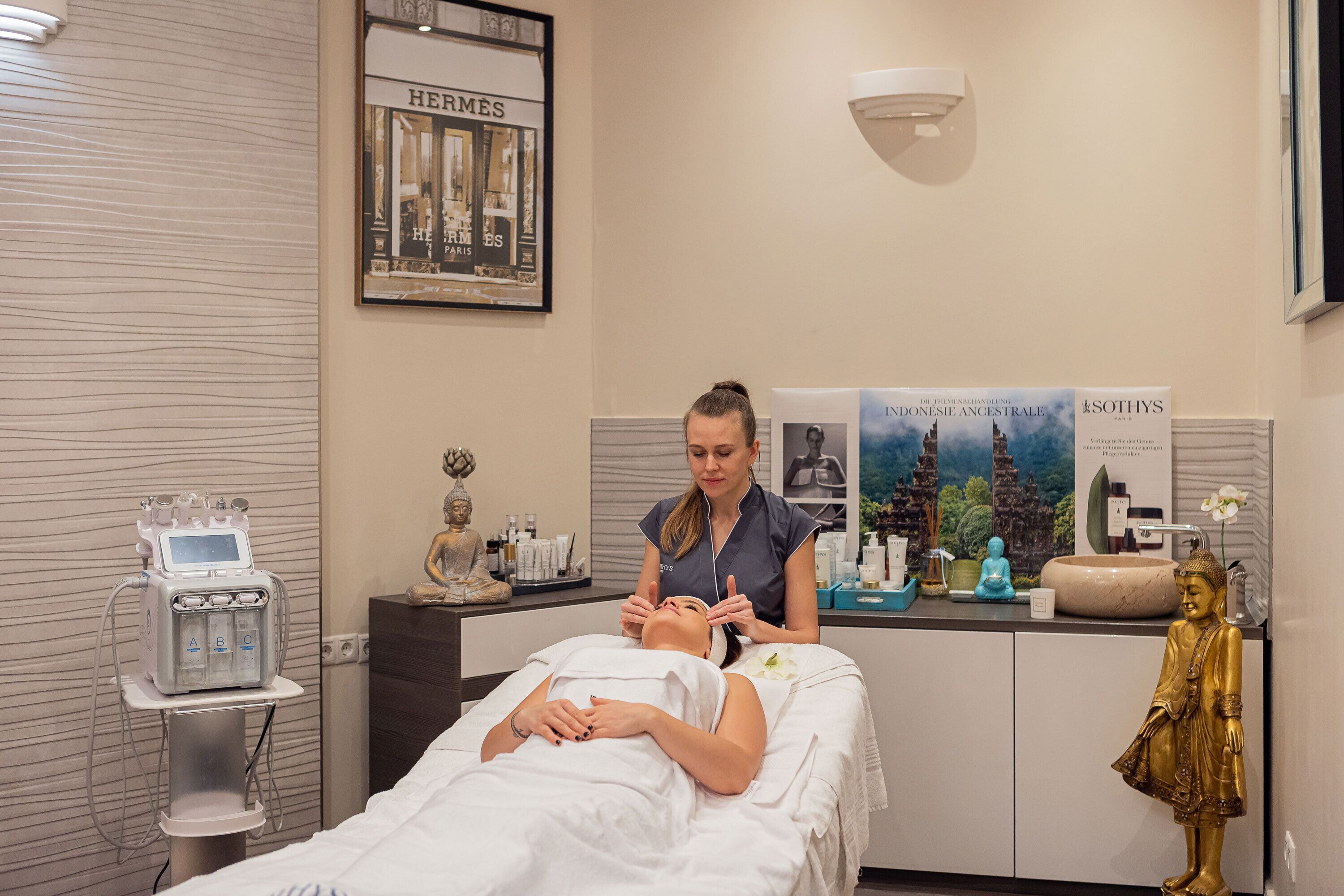 Une salle de soins avec une table de massage, un(e) massothérapeute, un appareil pour les soins du visage et une statue de Bouddha.