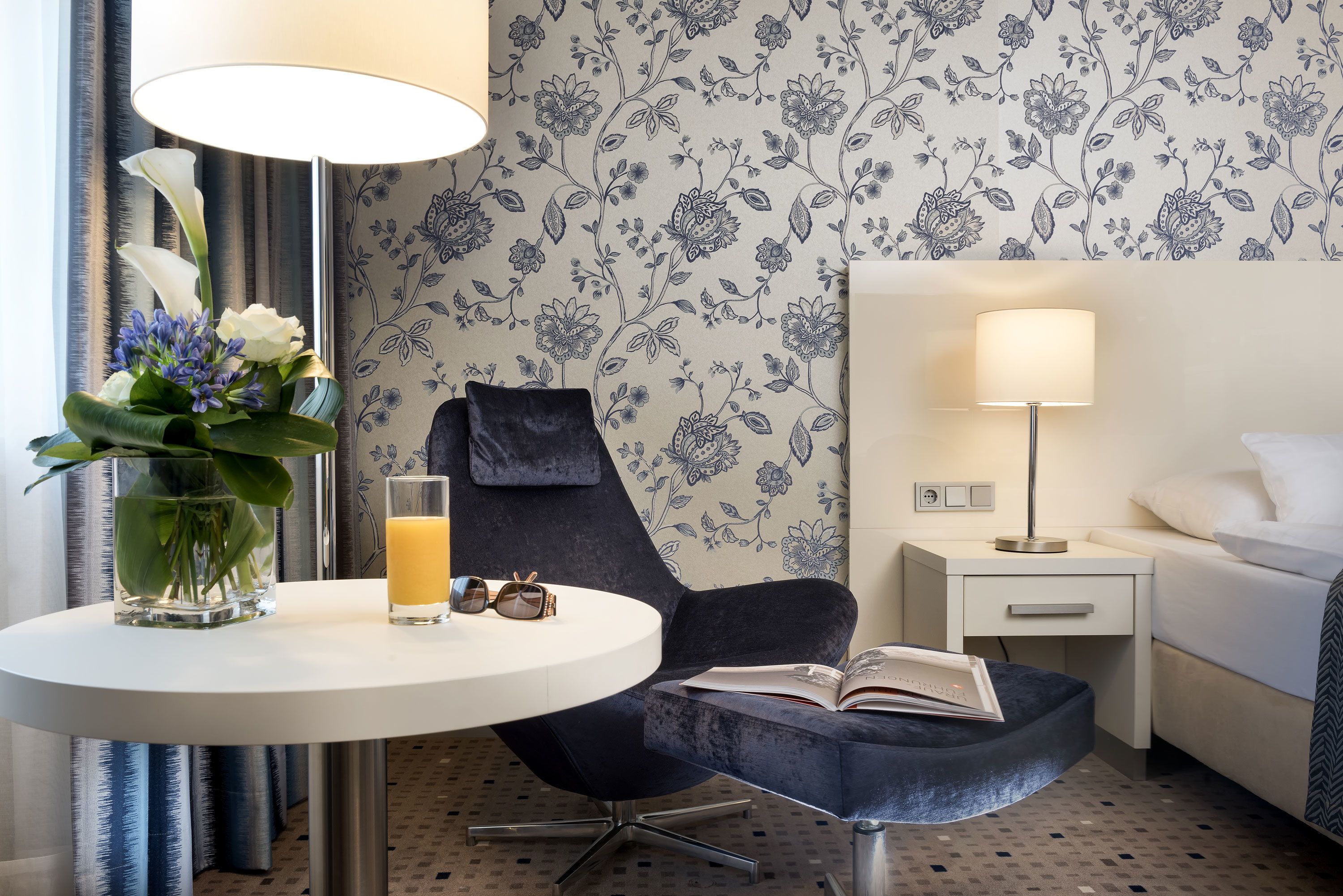 A hotel room with a round table, a dark armchair, a bedside table with a lamp, a bed, and a floral wallpaper.