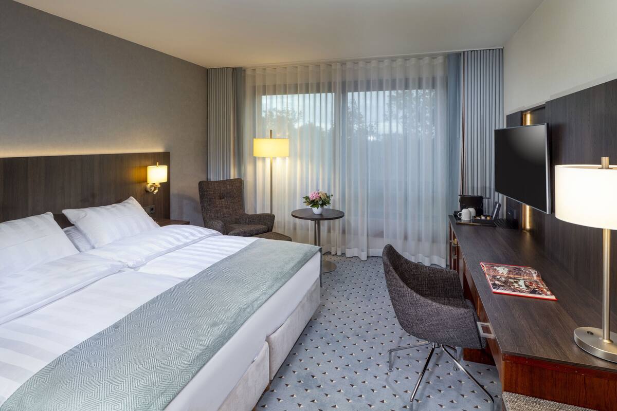 Ein Hotelzimmer mit einem großen Bett, einem Schreibtisch mit Fernseher, einem Sessel, einem kleinen Tisch mit Lampe und einem Fenster mit Vorhängen.