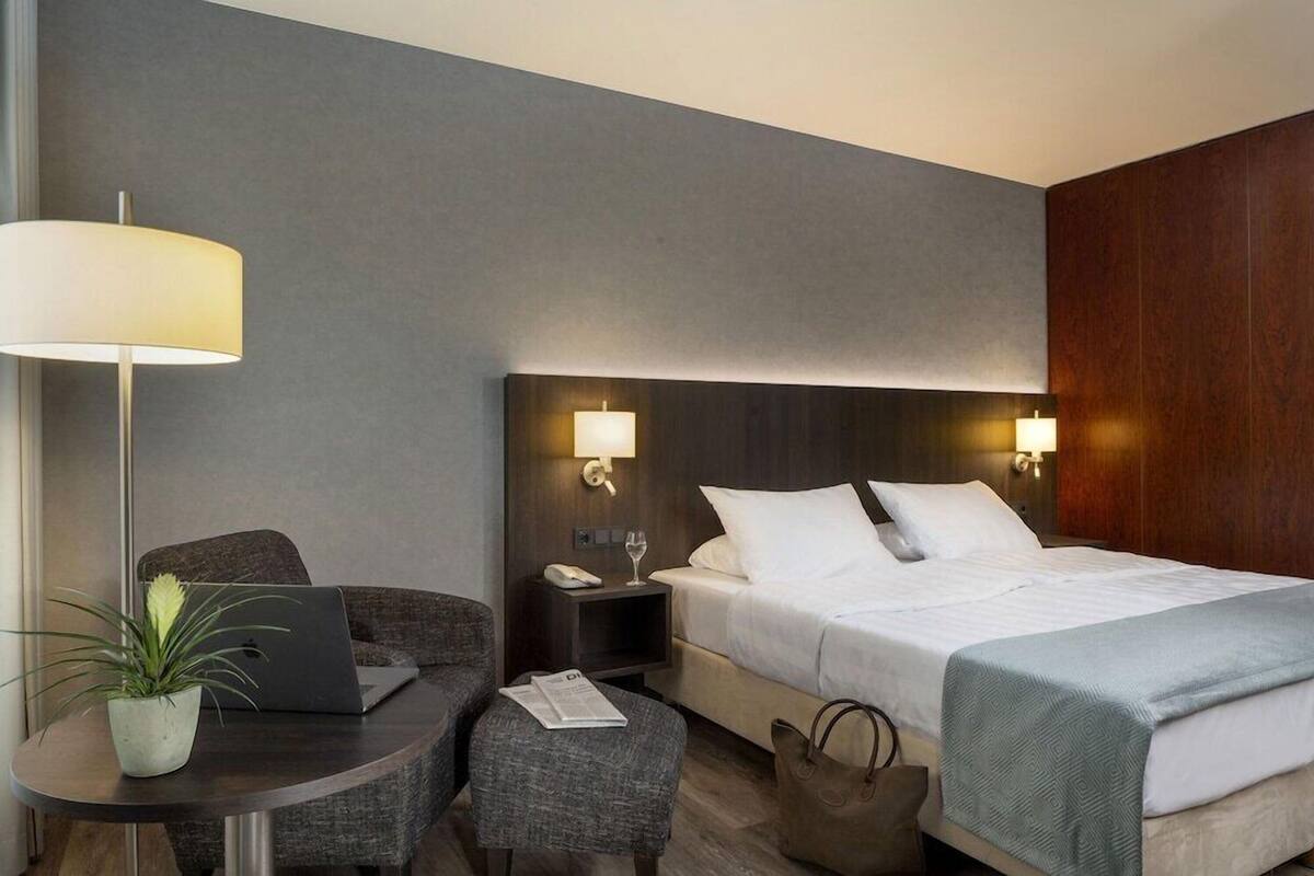 Ein Hotelzimmer mit Bett, Nachttischlampen, einem Schreibtisch mit Laptop, einem Stuhl, einem kleinen Tisch mit Pflanze und einer Handtasche auf dem Boden.