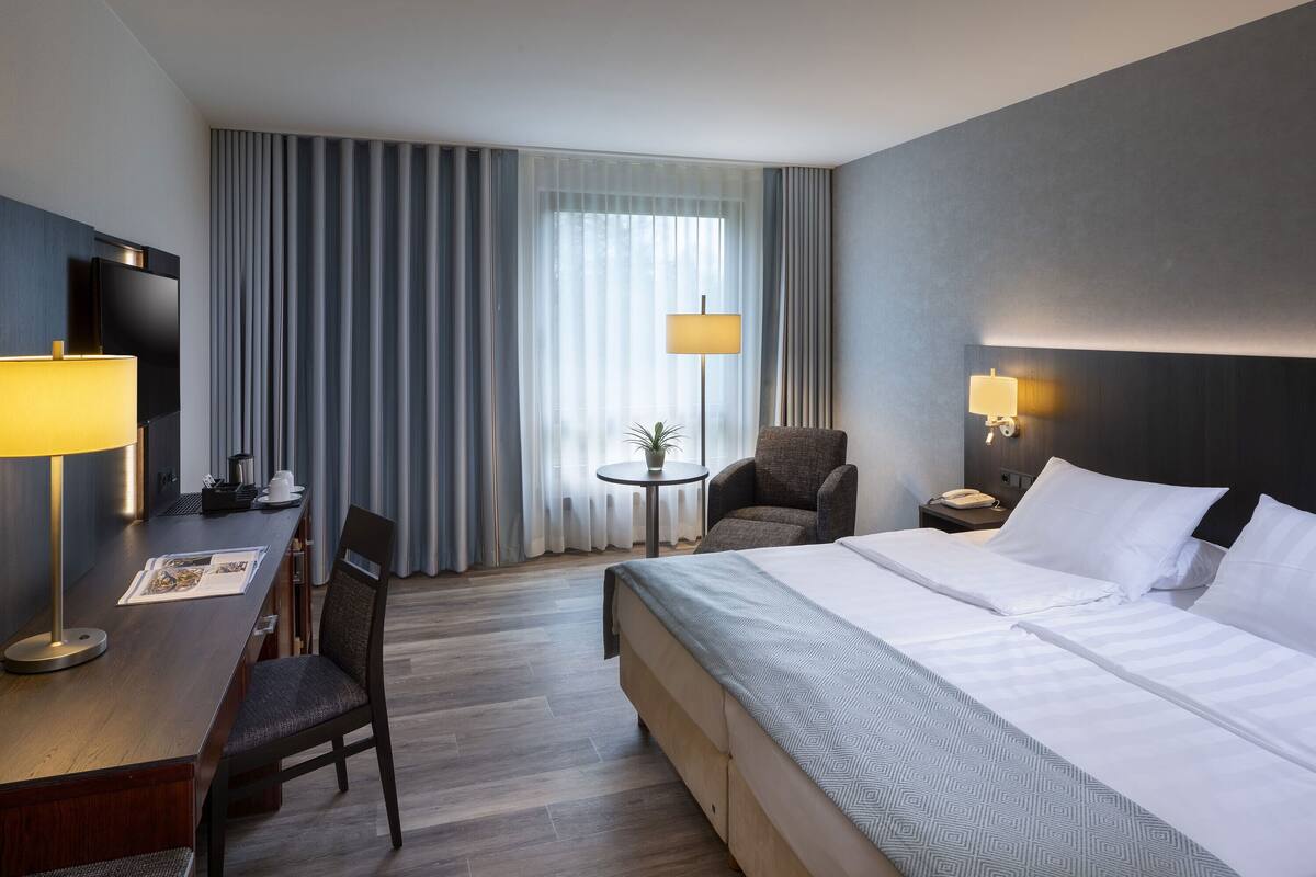 Ein Hotelzimmer mit einem großen Bett, einem Schreibtisch mit Lampe, einem Sessel, einem kleinen Tisch mit Pflanze und einem Fernseher.