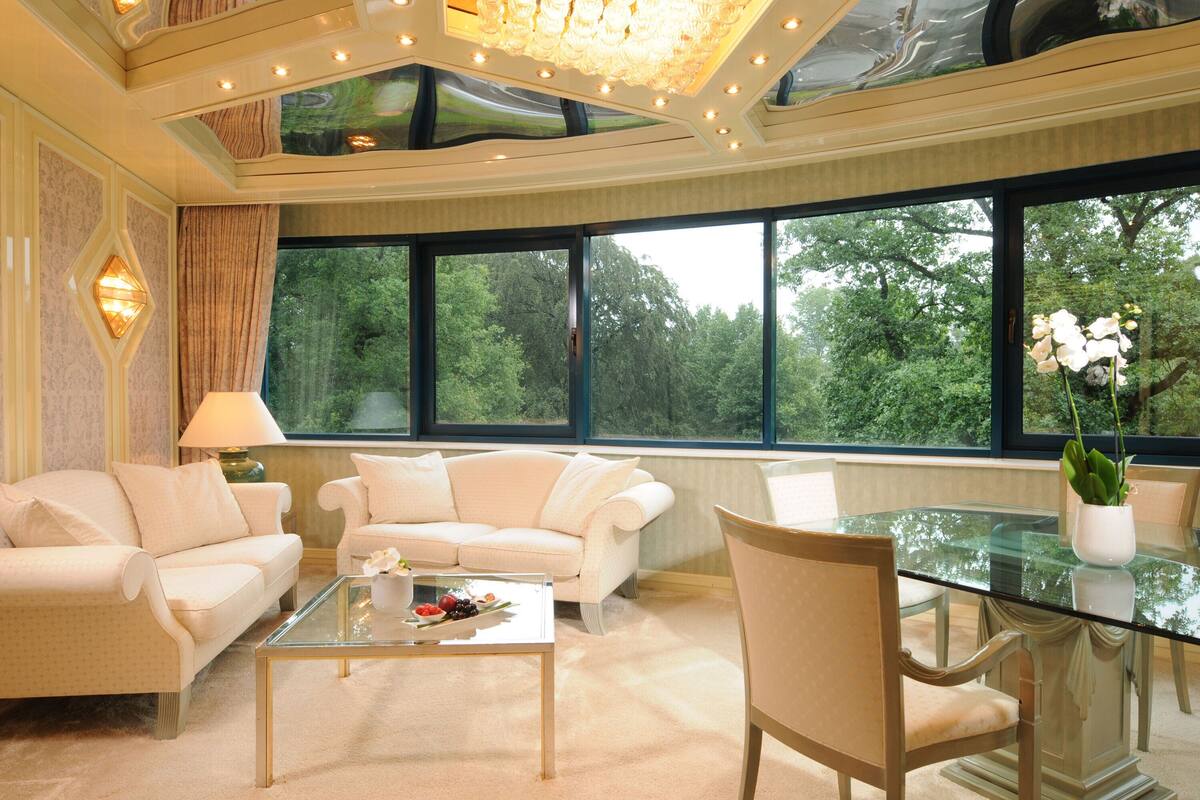 Ein modernes Wohnzimmer mit einem Glastisch, zwei weißen Sofas, einem Esstisch mit Stühlen und großen Fenstern mit Blick auf Bäume.