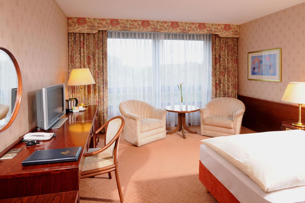 Ein Hotelzimmer mit einem Bett, zwei Sesseln, einem kleinen runden Tisch, einem Schreibtisch mit Fernseher und einem großen Spiegel.