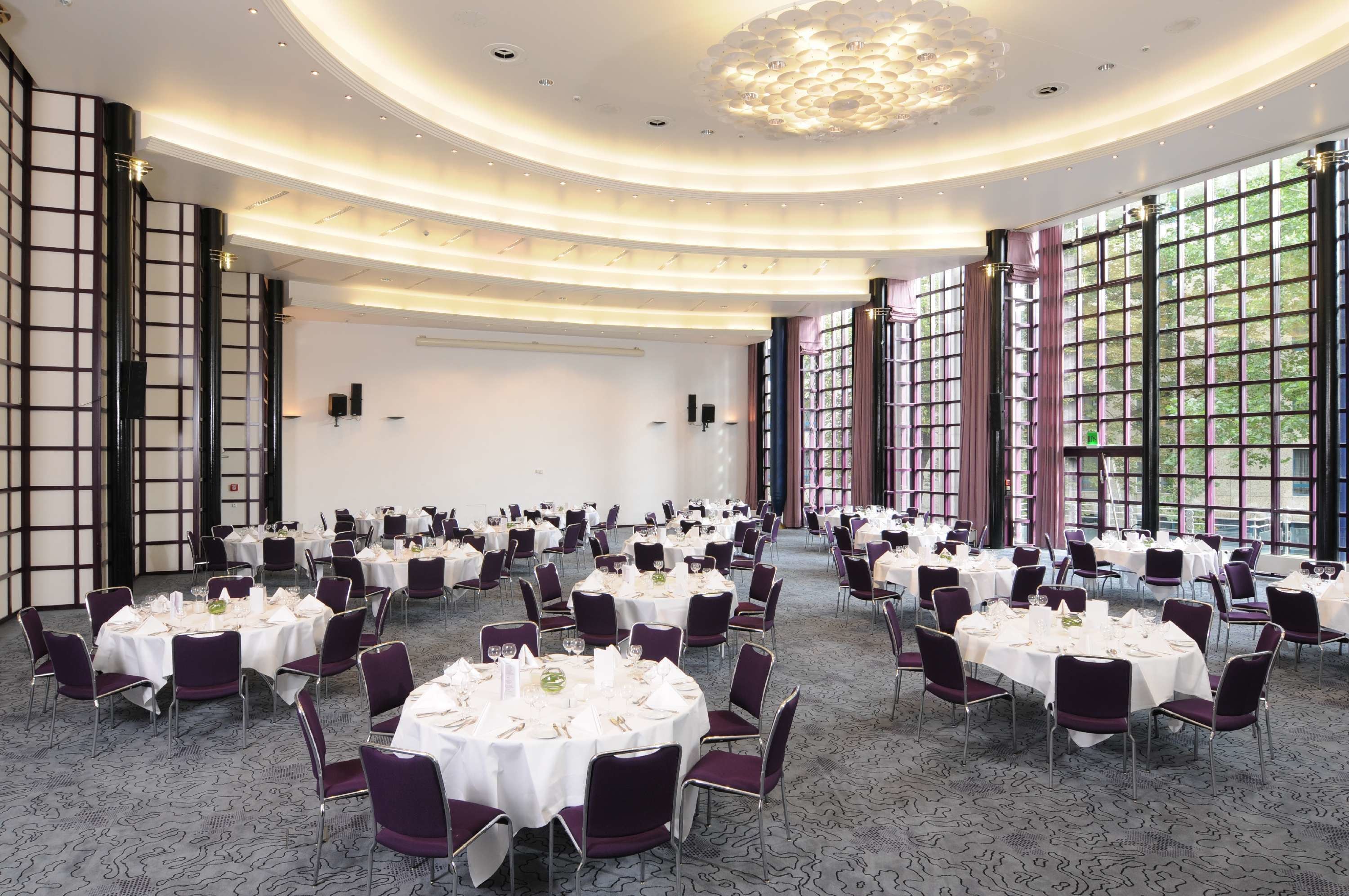 A large banquet hall with round tables set for dining, featuring a modern chandelier and large windows.